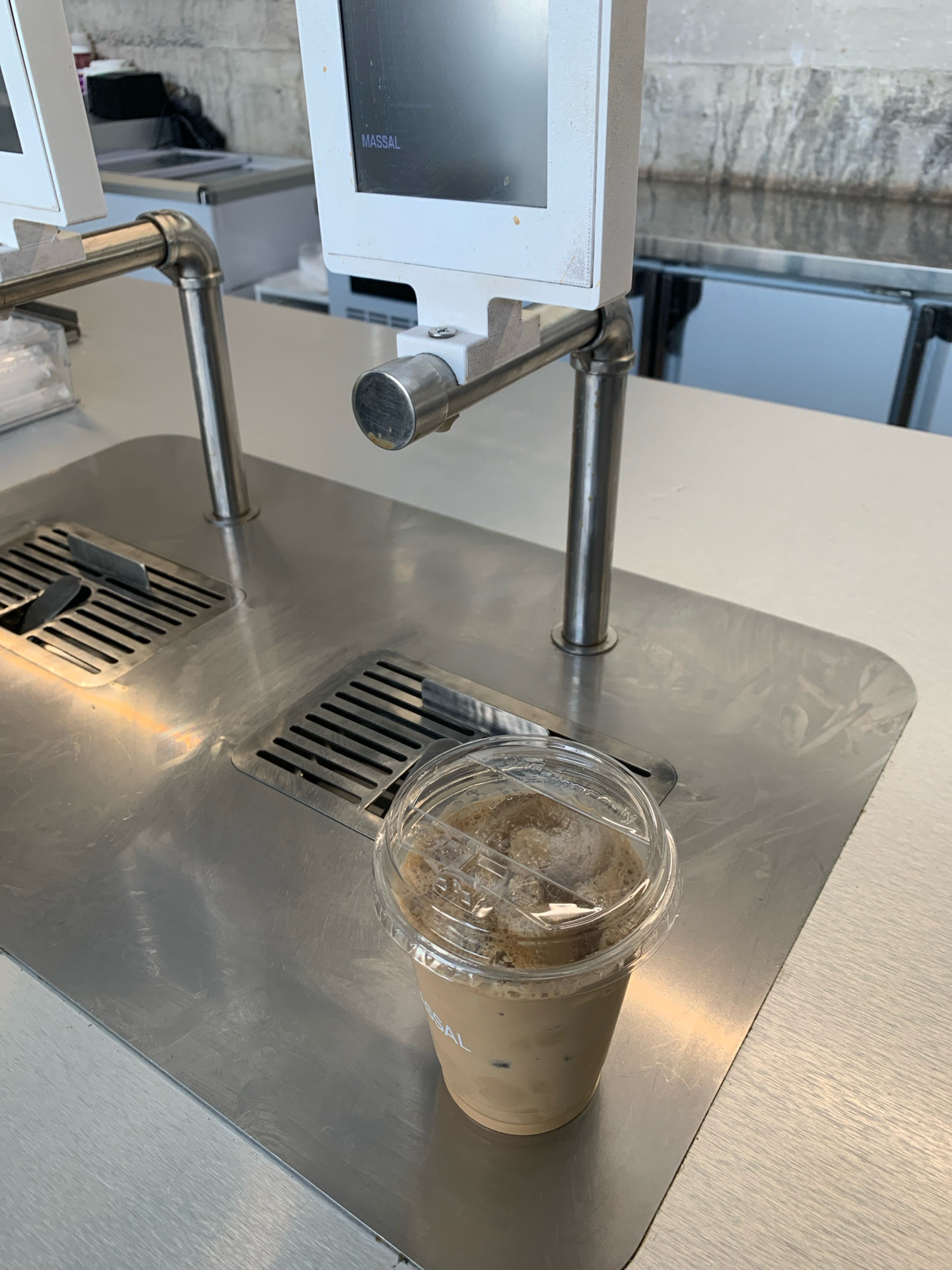 A clear plastic cup filled with an iced coffee drink rests on a stainless steel counter. The cup has a clear plastic lid with a sipping hole. Behind the cup are two stainless steel grates inset into the counter. A silver metal arm extends upwards from the counter, supporting a white device with a dark screen. Text reading "MASSAL" is visible on the screen. The background shows a concrete wall and reflective surfaces that appear to be part of a commercial establishment, possibly a cafe or restaurant. There are no people visible in the image. The scene is clean and modern. The location is not specified in the image itself.
