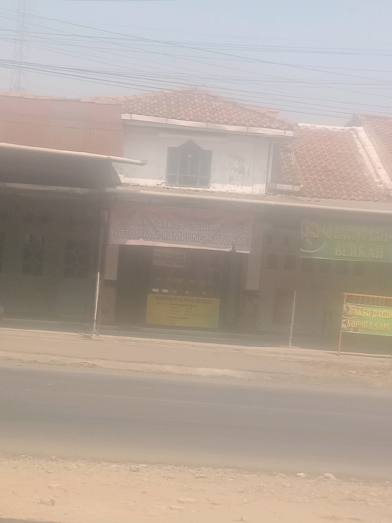 The image shows a street scene in Lebaksiu, Indonesia, with a hazy atmosphere. In the foreground, a paved road is visible, with a sandy shoulder on the right. Behind the road, a building with a tiled roof and a covered storefront stands. Several signs are attached to the storefront. One sign, partially obscured by dust, reads "WM. H. BARKAH - SEDIA SOP IGA SOTO AYAM KAMPUNG, SOTO SAPI". Another sign on the right appears to say "BAKSO DAGING SAPI BERKAH". A third sign, to the far right, has text that is difficult to read but seems to include "BAKSO DAGIN SOP IGA SAPI". The building has windows and appears to be a food establishment. In the background, power lines and a tower are visible against a bright sky. There are no people or vehicles clearly identifiable in the scene. The overall impression is one of a roadside eatery on a dusty day.