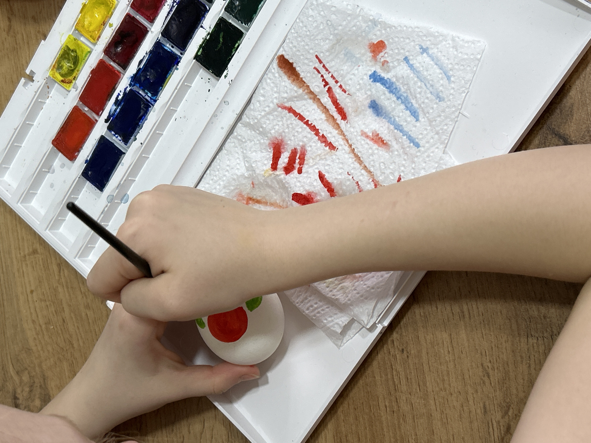 A person is painting an egg with watercolors. The egg, decorated with a red circle and green leaves, is held in one hand while the other hand holds a paintbrush. A watercolor palette with various colors and a paper towel with paint streaks sit next to the egg on a white tray. The background is a wood-patterned surface. The location is not discernible from the image.