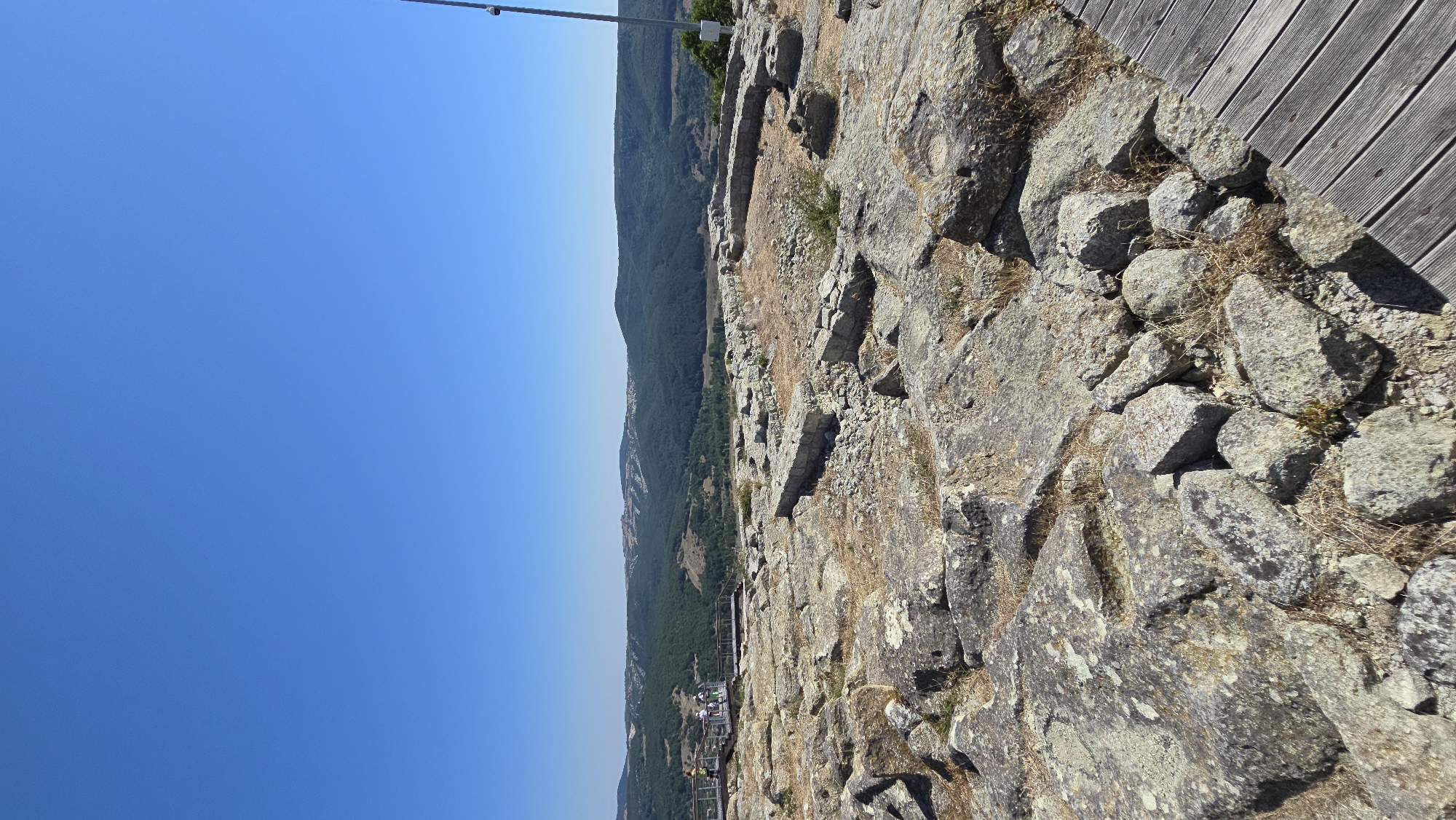 The image shows a panoramic view of an ancient archaeological site under a clear blue sky. The foreground is dominated by weathered, rough-hewn rocks, some of which appear to be remnants of ancient structures, possibly walls or foundations. Dry grass and sparse vegetation are scattered among the rocks. To the left, a wooden boardwalk with metal railings is visible, on which several people are standing and observing the landscape. In the midground and background, rolling hills covered in dense green forest stretch towards the horizon. A small town or settlement with red-tiled roofs is visible nestled in the hills in the distance. The site is located in the vicinity of Chiflik, Bulgaria, and is part of a larger historical or archaeological preserve. The overall scene suggests a sunny day with excellent visibility, ideal for outdoor exploration and sightseeing.
