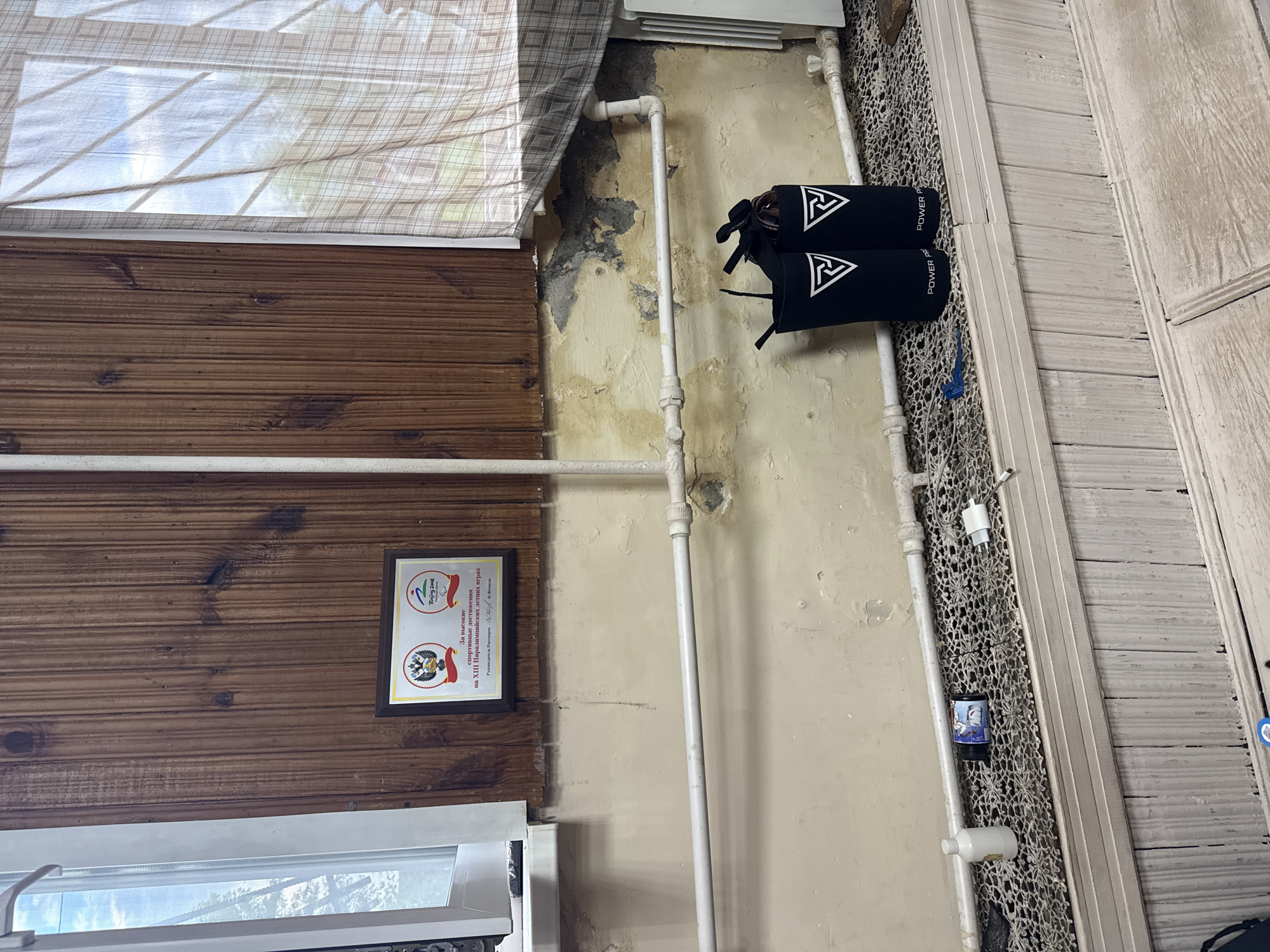 This is a candid, medium shot of an indoor scene, likely a room in Kazan, Russia. The camera is positioned at a slight downward angle, capturing a corner of the room where the wall meets the floor. The wall is a pale, textured yellow, showing signs of wear and peeling paint, with a darker patch near the ceiling, possibly water damage. A white pipe runs vertically along the wall.
To the left, a section of the wall is covered in dark wooden paneling, with visible wood grain and knots. A framed certificate hangs on the wooden paneling, featuring the Beijing 2008 Paralympic Games logo and text in Russian. The text translates to "For high sports achievements at the XIII Summer Paralympic Games" and mentions "Head of Rosport."
Above the wooden paneling, a plaid curtain or blind is visible, casting shadows from what appears to be a window with multiple panes. The curtain is a light beige and white checkered pattern.
In the center of the image, two black sleeves or guards are hanging from the white pipe. They have a white triangular logo with three arrows pointing inward, and the word "POWER" is printed in white text.
To the right of the pipe and sleeves, a decorative white lattice or mesh covers a portion of the wall. A white power adapter is plugged into something behind the lattice, and a black cylindrical object with a label featuring a drawing of a sailing boat is also visible. Further to the right, a light-colored wooden door or cabinet with vertical panels is seen. The overall impression is of a lived-in space with practical elements and a touch of personal history. There are no people visible in the image, and the lighting suggests it is daytime.