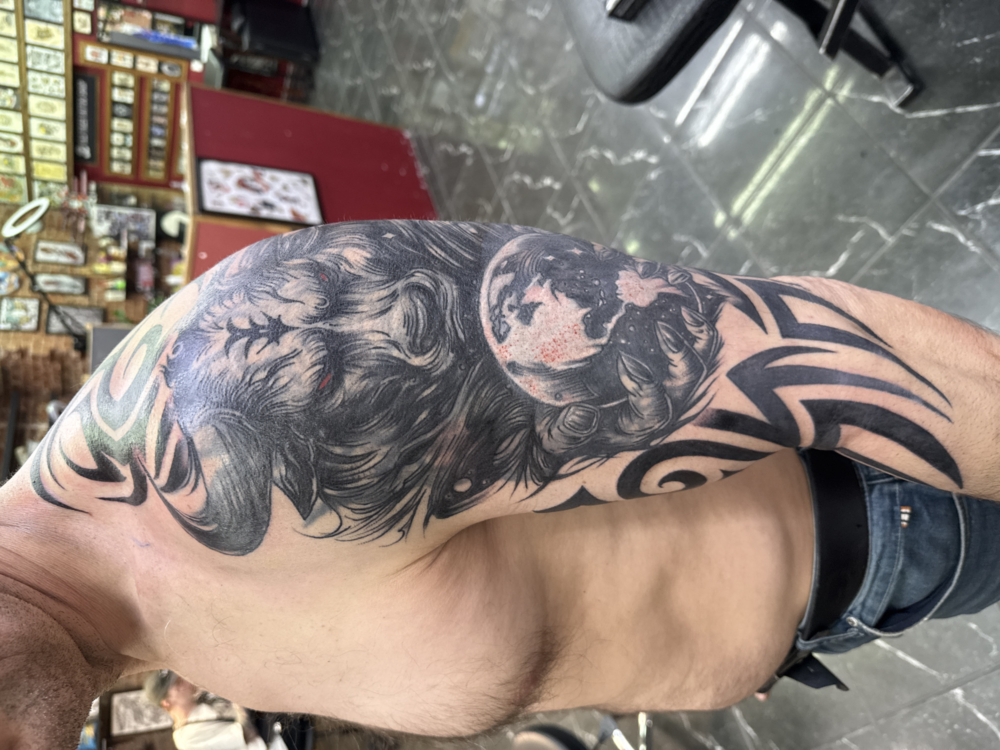 A man's upper left arm and shoulder are displayed, featuring a recently applied black and gray tattoo that extends from the shoulder down the upper arm. The tattoo design includes a depiction of a wolf's head with red-tinged eyes, an image of Earth, and tribal patterns. The man is shirtless, wearing blue jeans and a black belt. The background suggests a tattoo parlor, with tattoo designs displayed on the walls and tattoo equipment visible. The floor is tiled with a gray diamond pattern. There is no evidence to suggest that the image was taken in Moscow, Russia.