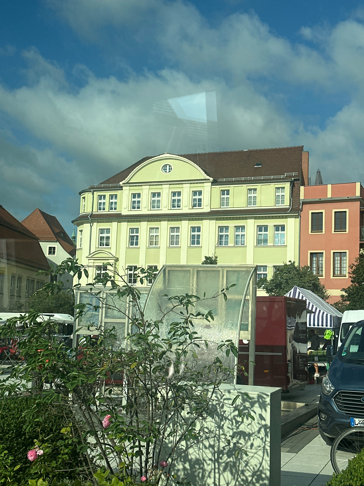 The image shows an outdoor scene in Bautzen, Germany, likely a market or event. In the foreground, a rose bush with pink blooms is partially obscuring a transparent structure. Behind the bush, a red vendor stall is visible, along with a striped awning and a dark blue van. The background features several buildings: a light green building with numerous windows, a peach-colored building with a darker trim, and a brown-roofed building with a steep gable. The sky is blue with scattered clouds, and reflections are visible on the transparent structure in the foreground.
