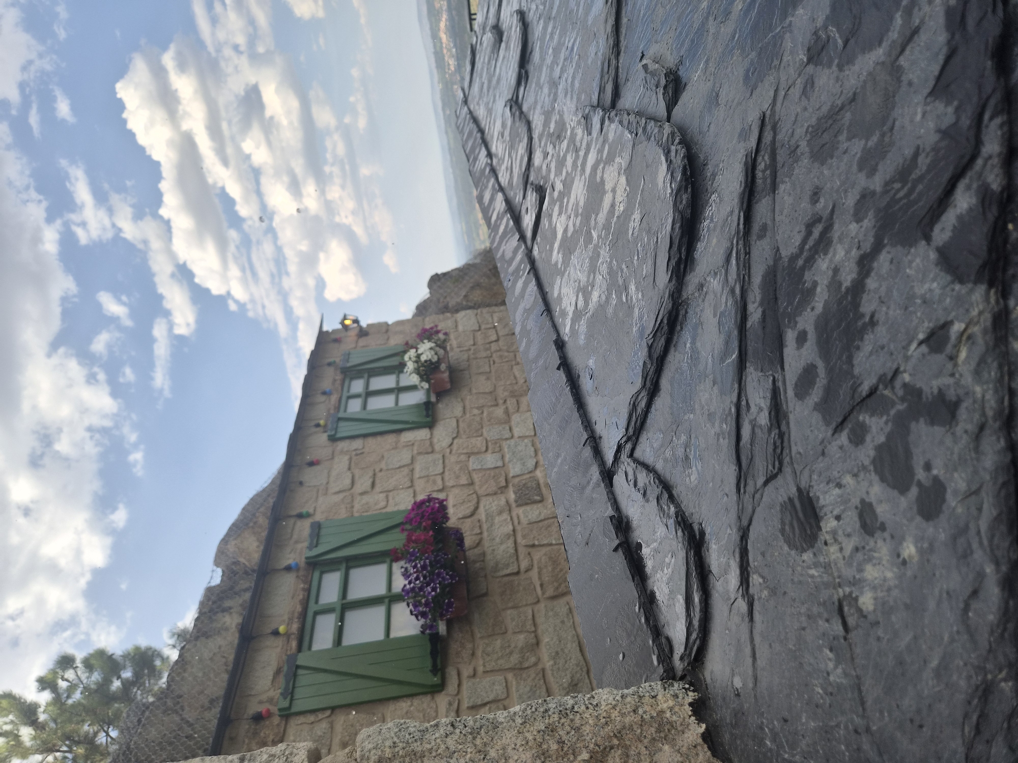 The image shows a close-up view of a dark gray, slate roof with visible water droplets, indicating it may have recently rained or is damp. The roof is textured with overlapping, irregular-shaped slates. In the background, a stone building with two green-shuttered windows is visible. One window has a planter filled with purple and pink flowers, and the other has a planter with white flowers. A string of colorful lights is draped across the top of the building. The building appears to be situated on a hillside or rock formation. Beyond the building, a landscape with rolling hills and a distant town can be seen under a blue sky with scattered white clouds. The overall scene suggests a rustic, possibly mountainous, location in Seia, Portugal. No people are present in the image.