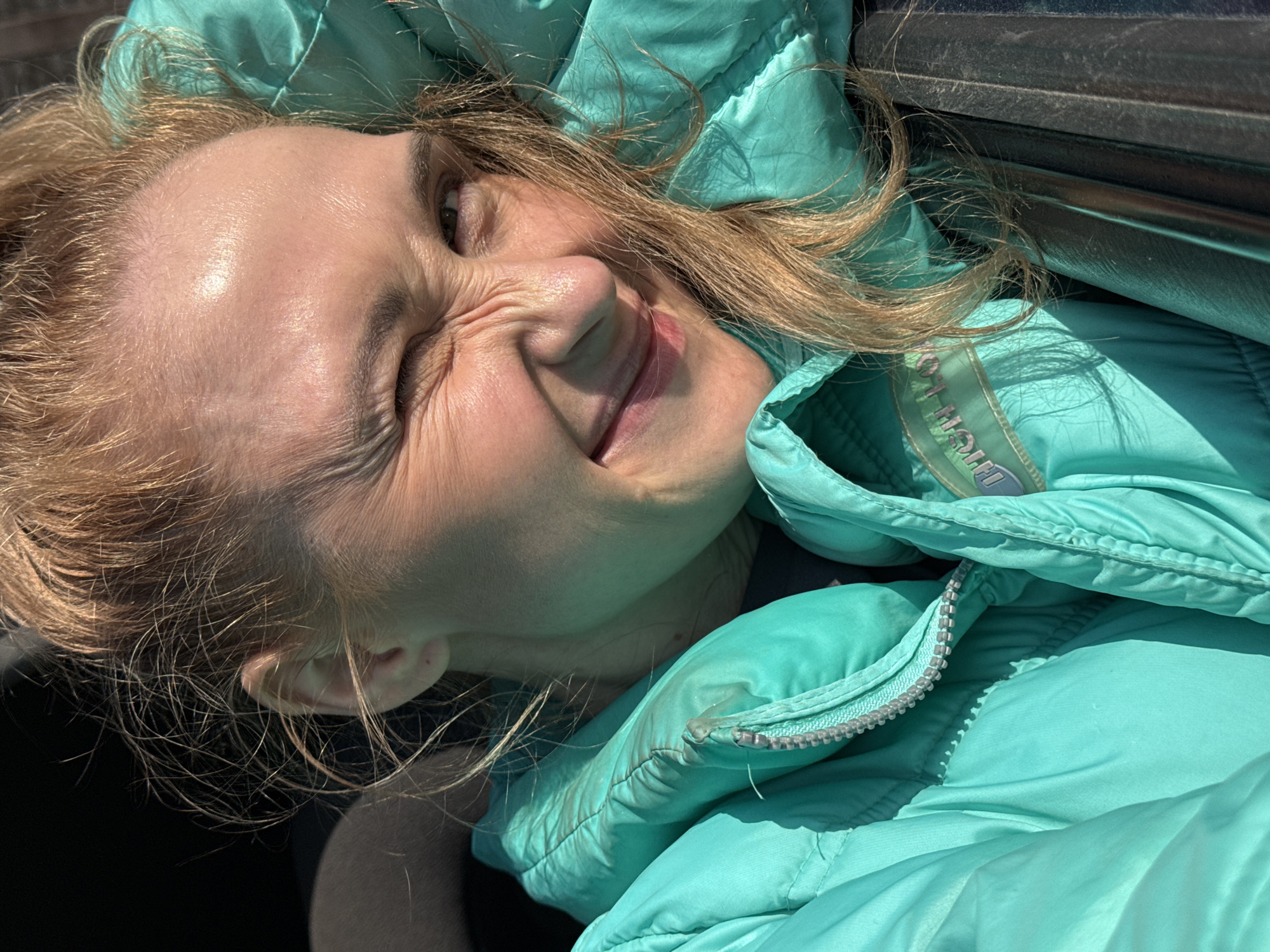 A woman with blonde hair and a light complexion is lying on her side, likely outside, as she is wearing a light-blue padded jacket. Her face is contorted into a squint with one eye closed, suggesting she is either smiling intensely or reacting to bright light. The location, Chaikovsky, Russia, is not discernible from the image.