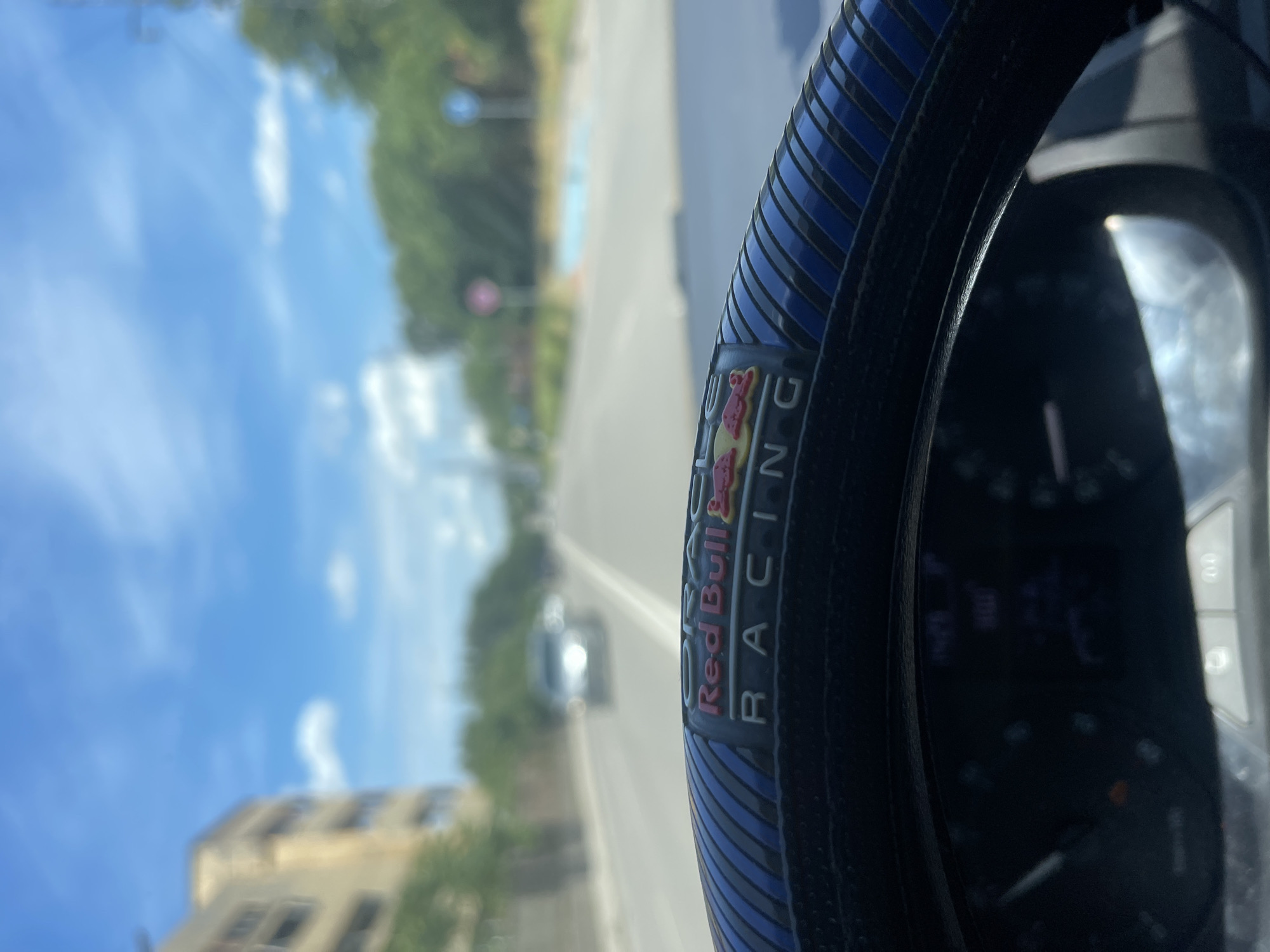 The image is a first-person perspective from inside a car, looking forward through the windshield. The foreground is dominated by a steering wheel with a blue and black cover, featuring a "Red Bull RACING" emblem. The steering wheel is slightly out of focus, as is the rest of the scene.
The view ahead shows a paved road with lane markings. There is a car in the distance, driving in the same direction. On the left side of the road, there is a building that appears to be residential or office, also out of focus. On the right side of the road, there are trees and a sidewalk. In the background, the sky is blue with scattered white clouds.
There are several circular signs visible on the right side of the road in the distance; one appears to be red and another blue, possibly indicating traffic regulations.
The specific location is identified as Ruse, Bulgaria, based on external information about the image's origin. The overall impression is a snapshot of a drive on a sunny day in an urban or suburban environment in Ruse. No people are clearly visible.