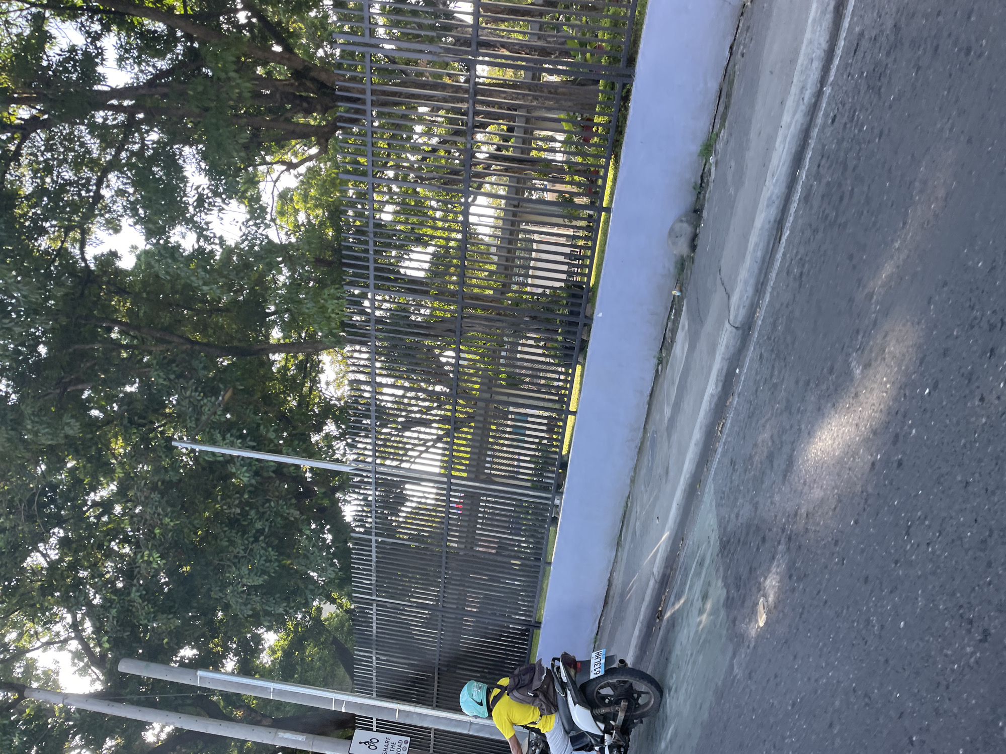 This image captures an outdoor scene featuring a motorcyclist on a paved road, adjacent to a secured area with dense vegetation. The photograph appears to be taken during daylight hours, evidenced by bright sunlight filtering through tree canopies and casting shadows on the asphalt.
In the mid-ground, a person is seen from the rear, riding a white and black motorcycle. The rider is wearing a bright yellow short-sleeved shirt, light-colored pants, and a light blue or teal full-face helmet. A dark brown backpack is strapped to the rider's back. The motorcycle's rear license plate, "613LHH," is visible.
To the right of the motorcycle, a light blue-gray concrete wall or curb separates the road from a strip of green grass, behind which stands a tall, dark gray metal fence composed of vertical bars. The fence runs horizontally across the frame, partially obscuring the background. To the left, a white rectangular road sign featuring a black bicycle icon and the text "SHARE THE ROAD" is mounted on a grey pole. Several other grey utility poles are present to the left of the frame, some with visible wiring.
The background is dominated by large trees with abundant green foliage, through which sunlight penetrates, creating bright patches. Partially visible behind the fence and trees are hints of light-colored structures. The road surface in the foreground shows various textures and lighter patches, possibly sunlight reflections or debris.
This scene is located within Davao City, Philippines.