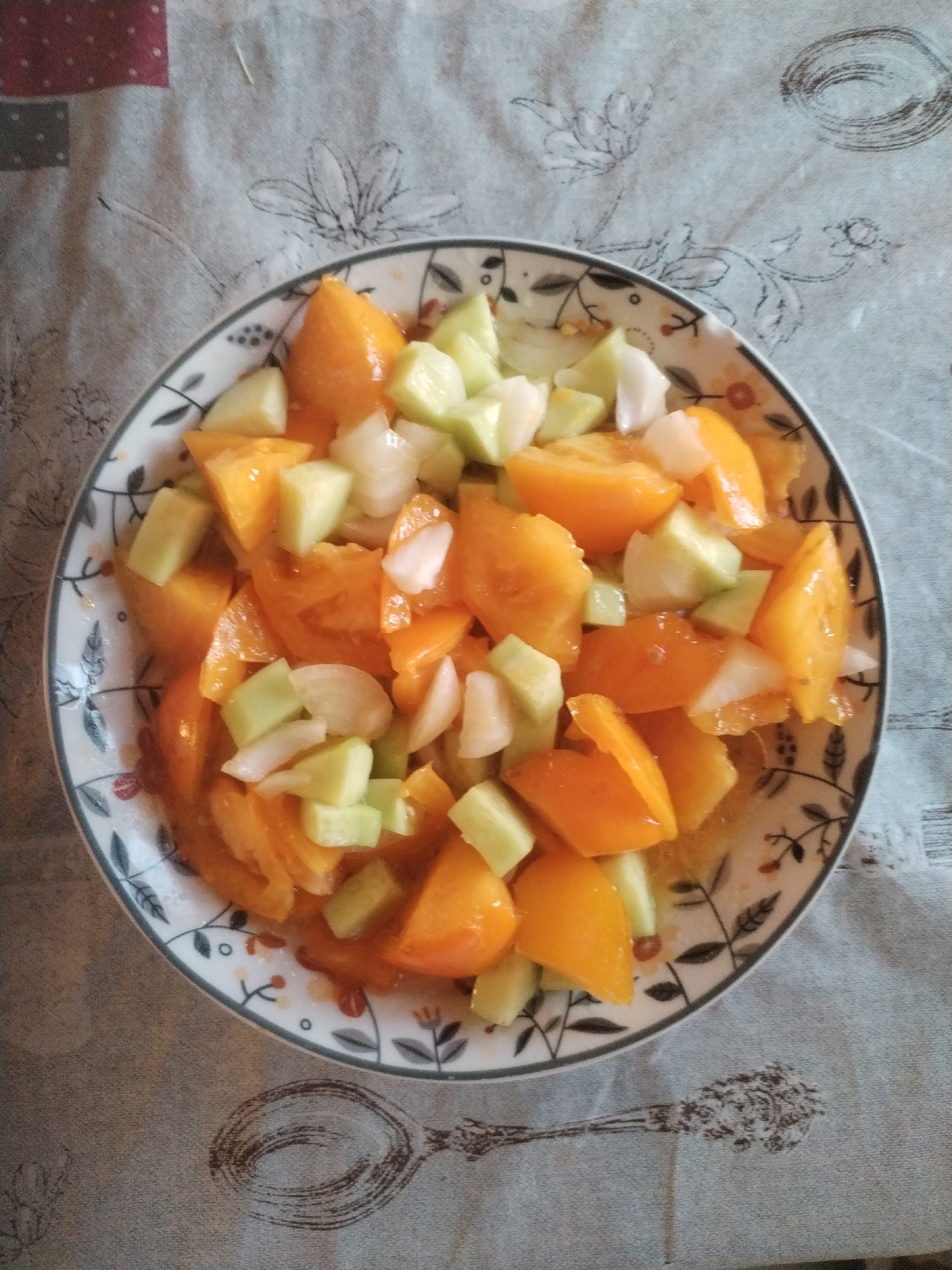 A close-up, top-down shot shows a bowl of chopped yellow tomatoes and cucumbers on a tablecloth with a floral pattern. The bowl is white with a black and gray decorative rim. The tomatoes are cut into bite-sized wedges, and the cucumbers are diced. The scene is lit from above, casting subtle shadows. The tablecloth is gray with a repeating pattern of black floral designs and outline sketches of utensils. The image does not provide any information about the city of Moara Domnească, Romania, or any people present.