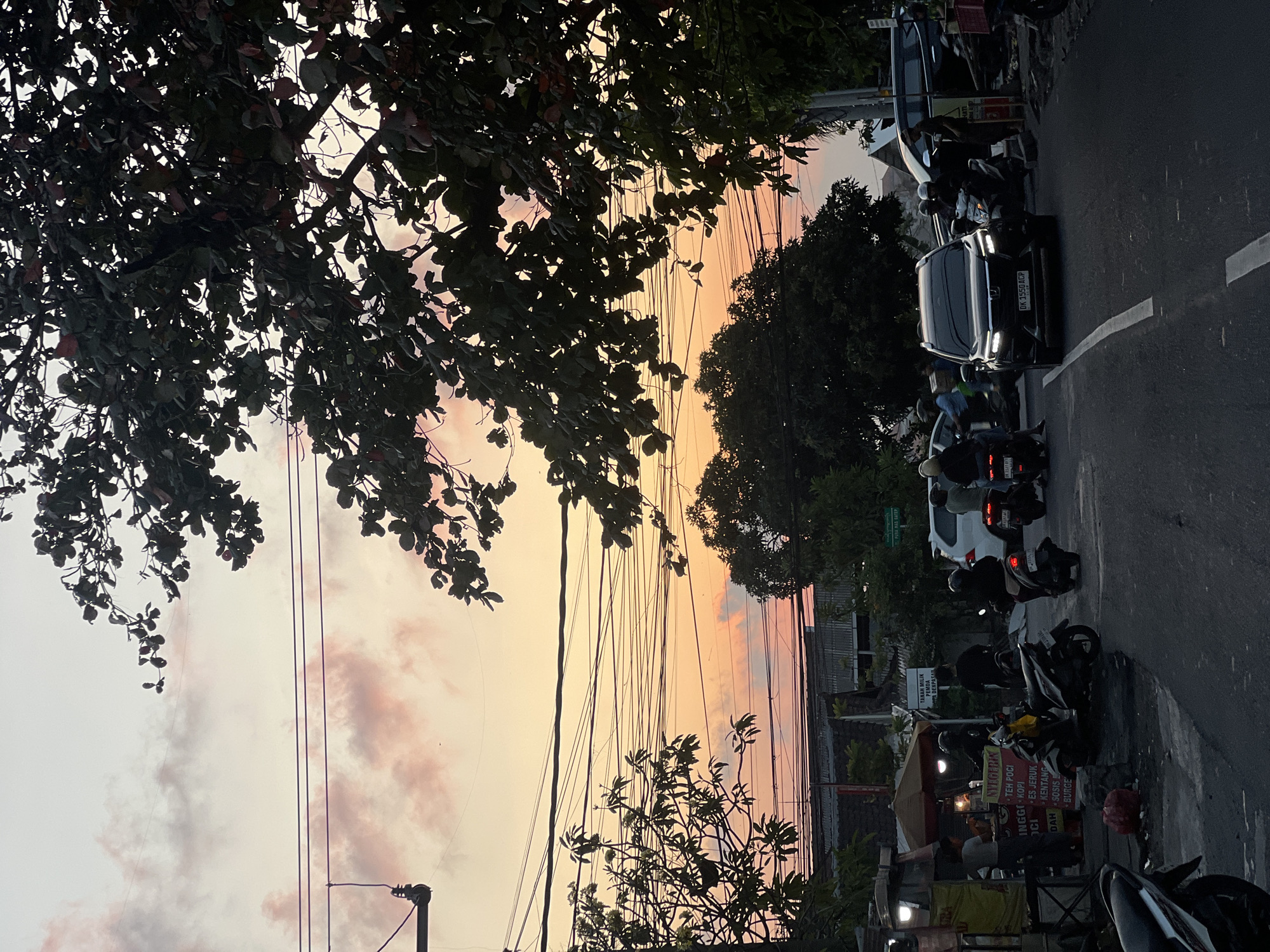 This is a medium shot of a street scene in Denpasar, Indonesia at dusk. The sky is a gradient of soft pink and orange, with scattered clouds. Overhead, a complex web of electrical wires crisscrosses the frame. In the foreground, the asphalt road is marked with a white dashed line. To the left of the road, a street food stall with a yellow awning is visible, with several people gathered around it, including one person standing behind the counter. Several motorcycles are parked or being ridden on the street. A black SUV with license plate DK 1550 ACP is driving towards the camera, its headlights on. To the right of the road, trees and vegetation line the street, with some buildings visible behind them. The overall atmosphere is that of a busy street at the end of the day, with the natural beauty of the sunset contrasting with the urban environment.