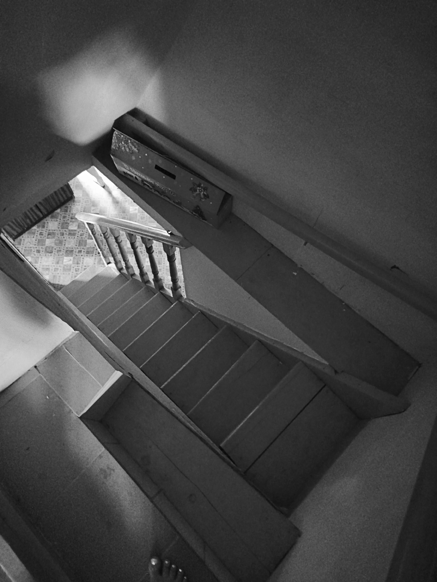 This forensic description pertains to an interior scene within a residential or multi-story building in Терновский округ, Ukraine.
The image is a high-angle, black and white photograph taken from the top of a staircase, looking downwards. The perspective is significantly tilted.
**Subjects and Objects:**
The primary subject is a multi-turn wooden staircase, consisting of multiple steps and a wooden balustrade on its left side (relative to the descent). The balustrade features turned wooden balusters and a handrail.
The flooring at the bottom of the visible staircase is patterned with a light-colored, repeating geometric tile or linoleum design.
On the immediate bottom edge of the frame, the toes of a human foot, with painted toenails, are visible, indicating the presence of the photographer.
On the upper right side of the image, against a wall or built-in structure, a decorative cardboard box is positioned horizontally. The box displays a pattern of stars and other motifs, possibly holiday-themed, and includes a dark, rectangular slot or opening on its visible side. Partial, indistinct text is also visible on the box.
The walls throughout the visible space are plain and exhibit textural variations, consistent with plaster or painted surfaces. Cracks are visible on the upper right wall segment.
A section of dark, horizontally striped fabric, possibly a curtain or blind, is partially visible in the upper left background, near the area where the staircase descends to the lower floor.
**Environment and Lighting:**
The environment is an indoor architectural space, characterized by domestic features. Lighting is provided by an unseen source, likely an opening or window at the lower level of the staircase, creating strong contrasts between brightly illuminated areas (e.g., the bottom floor, parts of the staircase) and deeply shadowed regions (e.g., under the stairs, sections of the walls). The scene is rendered in monochromatic tones, emphasizing light and shadow.