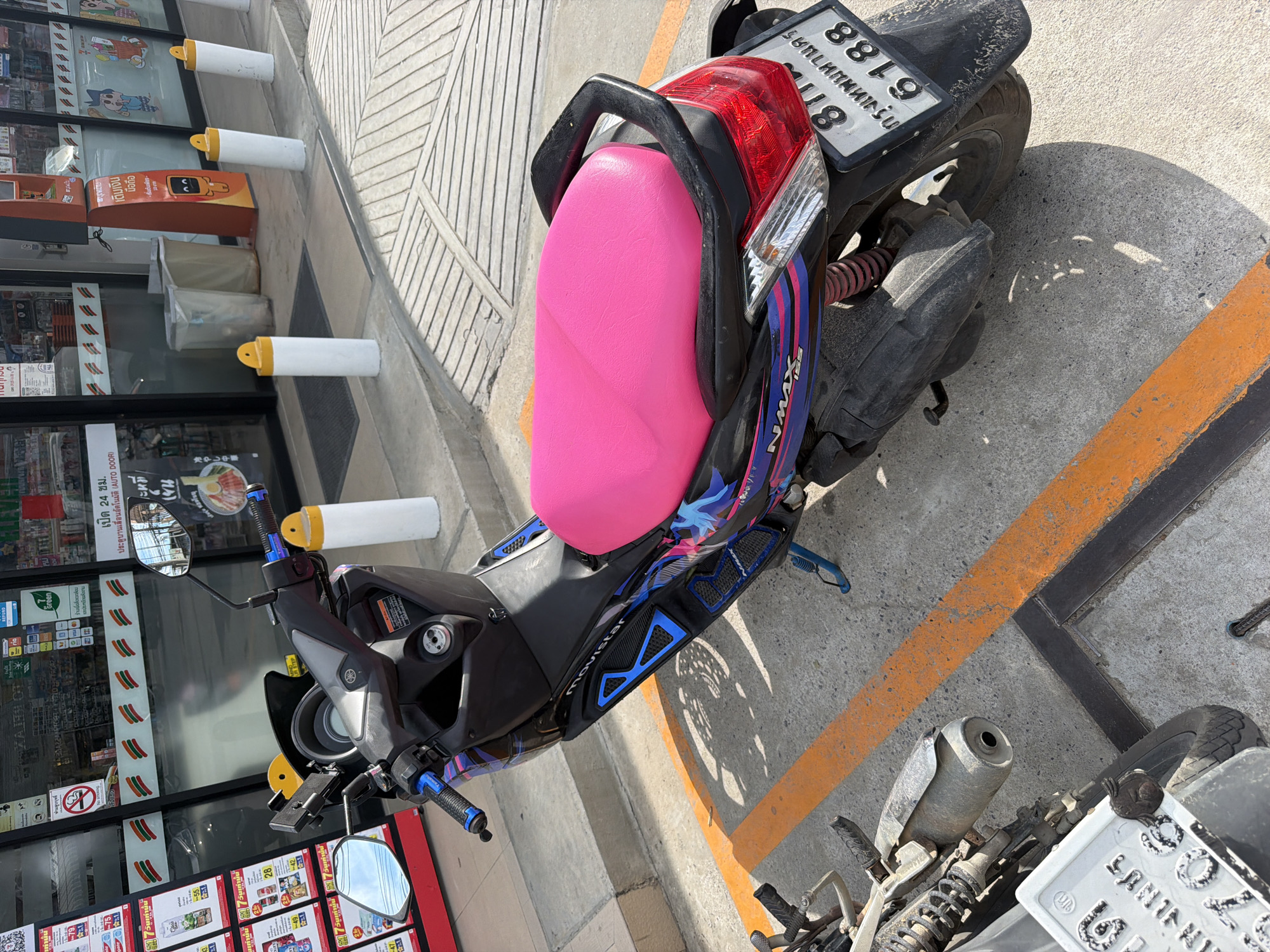 A Yamaha NMAX 155 scooter with a bright pink seat is parked on a concrete surface outside a 7-Eleven convenience store. The scooter has a black body with blue and pink graphic decals. A license plate with Thai characters and the number "811 6188" is visible on the rear. In the background, the storefront of the 7-Eleven is visible, with shelves of products displayed inside and signage in Thai. Several white bollards with yellow caps are positioned along the sidewalk in front of the store. Another motorcycle is partially visible in the foreground on the left side of the image. The scene appears to be a typical urban or commercial area in Baan Mae Nam, Thailand, likely during daylight hours due to the presence of shadows.
