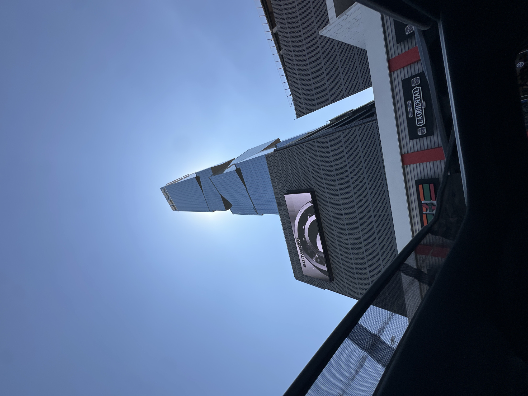 The image depicts a low-angle view of a modern urban development in San Bernardino Tlaxcalancingo, Mexico, under a clear, bright blue sky. A tall, slender skyscraper with an angular, stepped glass and panel facade dominates the center-right, with the sun's glare directly behind its upper section. Below the skyscraper, a multi-story building features a dark gray, textured facade. On this building, a large horizontal digital display exhibits "BIENVENID" text and abstract circular graphics in monochrome. Below the display, at a lower elevation, the facades of a "7-ELEVEN" store and an "Antigua Taquería LA ORIENTAL" restaurant, identified by its sign featuring beer mug logos, are visible against a backdrop of horizontal gray panels with red vertical accents. To the far right, another building with a similar dark gray facade is partially observed. The foreground reveals a dark interior frame and a mesh sunshade, indicating the photograph was taken from inside a vehicle, looking upward through a sunroof.