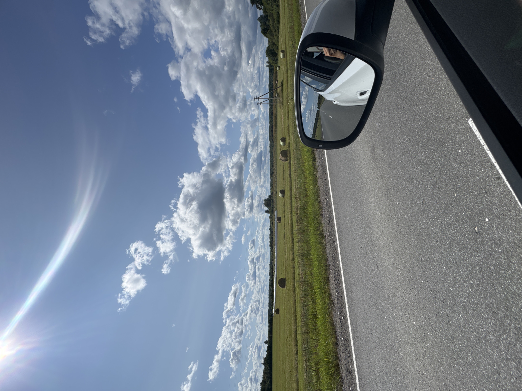 A side view from a moving car shows a bright, sunny day with a clear blue sky and scattered white clouds. The sun is visible in the upper left corner with a lens flare effect. A green field with numerous round hay bales stretches across the landscape, bordered by trees in the distance. A road with a white line runs in the foreground, with a portion of the car's exterior visible. The side mirror reflects the road ahead and the interior of the car, showing a person's arm and shoulder. There are power lines and utility poles in the background. The scene appears to be in a rural area of Spasskaya Polist', Russia.