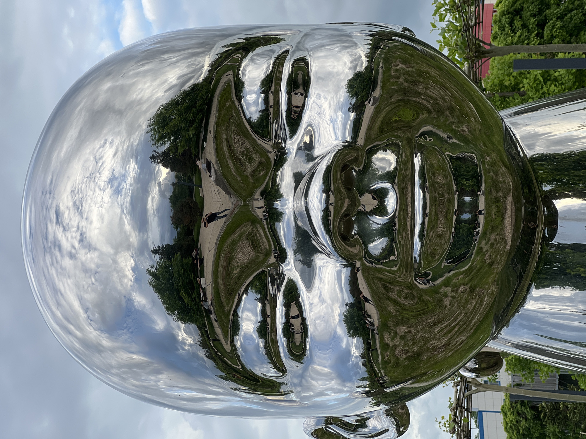 A shiny, reflective metal sculpture of a human head is the primary subject. The reflection in the sculpture shows a park with green trees, grass, and a cloudy sky. People are walking along the park's pathways. The image appears to have been taken in a park in the city of Krasnodar, Russia.