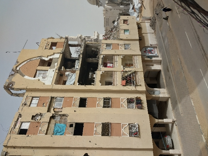 The image displays a street scene in Khan Yunis, Palestinian Territory, featuring a severely damaged multi-story building. The structure, characterized by a light-tan facade and ornate cream-colored balcony railings, exhibits extensive destruction. Large sections of the upper and right-side facades are missing, revealing destroyed interior rooms, exposed rebar, and concrete slabs. Numerous projectile impacts are visible across the remaining exterior walls. Most windows are shattered or entirely absent; a blue tarp hangs from a broken window on the left side of the building, while various clothes are draped over railings on multiple balconies.
At ground level, a paved two-lane road runs in front of the building, accompanied by a cobblestone sidewalk. A silver sedan is parked beneath an arched opening on the building's ground floor. Piles of rubble and construction debris are accumulated along the sidewalk and at the base of the structure. Two individuals are visible on the right side of the sidewalk: an adult figure in dark clothing, including a head covering, walks to the right, and a smaller figure, likely a child, sits or kneels on the ground nearby.
In the background to the right, further damaged structures are visible, including a building with a white dome and a minaret-like component, suggesting a mosque. Several white tents or temporary shelters are present amidst the debris in the distant background. The sky is uniformly pale grey.