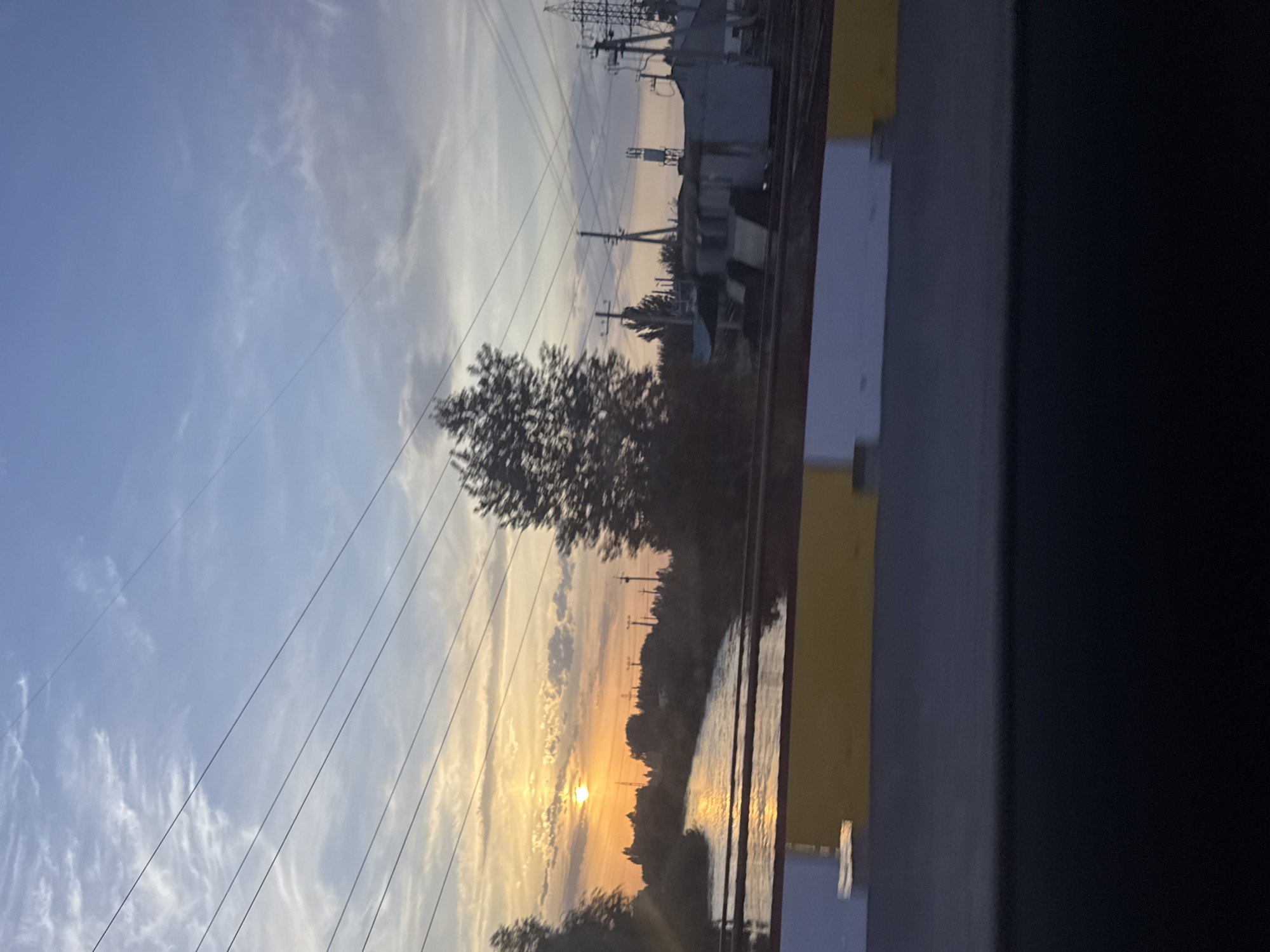 The image shows a scenic view of a river with the sun setting in the distance, casting a warm orange glow across the water. Power lines crisscross the sky, a common sight in many urban and rural areas. The riverbanks are lined with trees and vegetation, with a hint of industrial structures in the background. The foreground features a yellow and white barrier, possibly part of a bridge or road, suggesting the photo was taken from a moving vehicle. The sky is a mix of blue and pale orange, with wispy clouds that catch the last rays of sunlight. The overall impression is one of tranquility and natural beauty, juxtaposed with the man-made infrastructure. The image was taken in Lebedinovka, Kyrgyzstan.