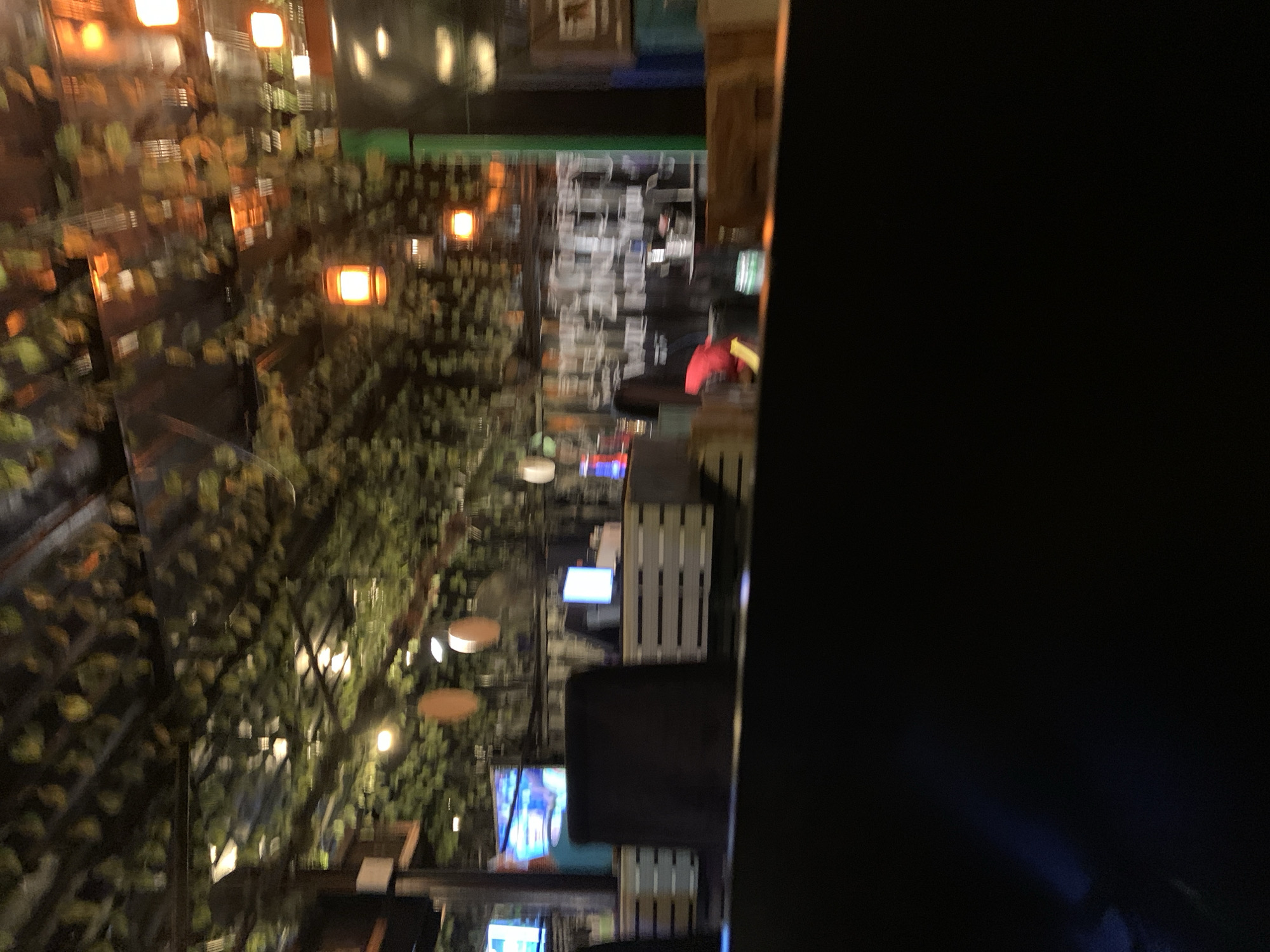 The image shows an indoor scene that appears to be a restaurant or bar, with a blurred, motion-affected aesthetic. The ceiling is decorated with artificial foliage and illuminated by numerous hanging light fixtures, some of which are square-shaped and emit a warm, orange glow. In the mid-ground, there are several tables or counter areas with what seem to be server stations. On one of these, a computer monitor displays a bright, blueish-white screen. Behind this, a dark, bulky object, possibly a chair or furniture, is visible. Further in the background, more shelving units and what could be bar equipment are present. The overall impression is that of a dimly lit, possibly lively, establishment, though the motion blur makes precise identification of objects and actions difficult. The location in Gura Top, Nigeria is not discernable from the visual information provided.
