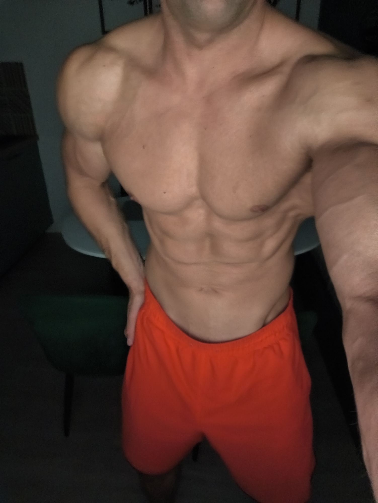 A shirtless man with visible musculature, wearing red shorts, stands in a dimly lit room. A green chair is visible to the left, along with a white table. The background is dark, obscuring details. The location is not discernible.