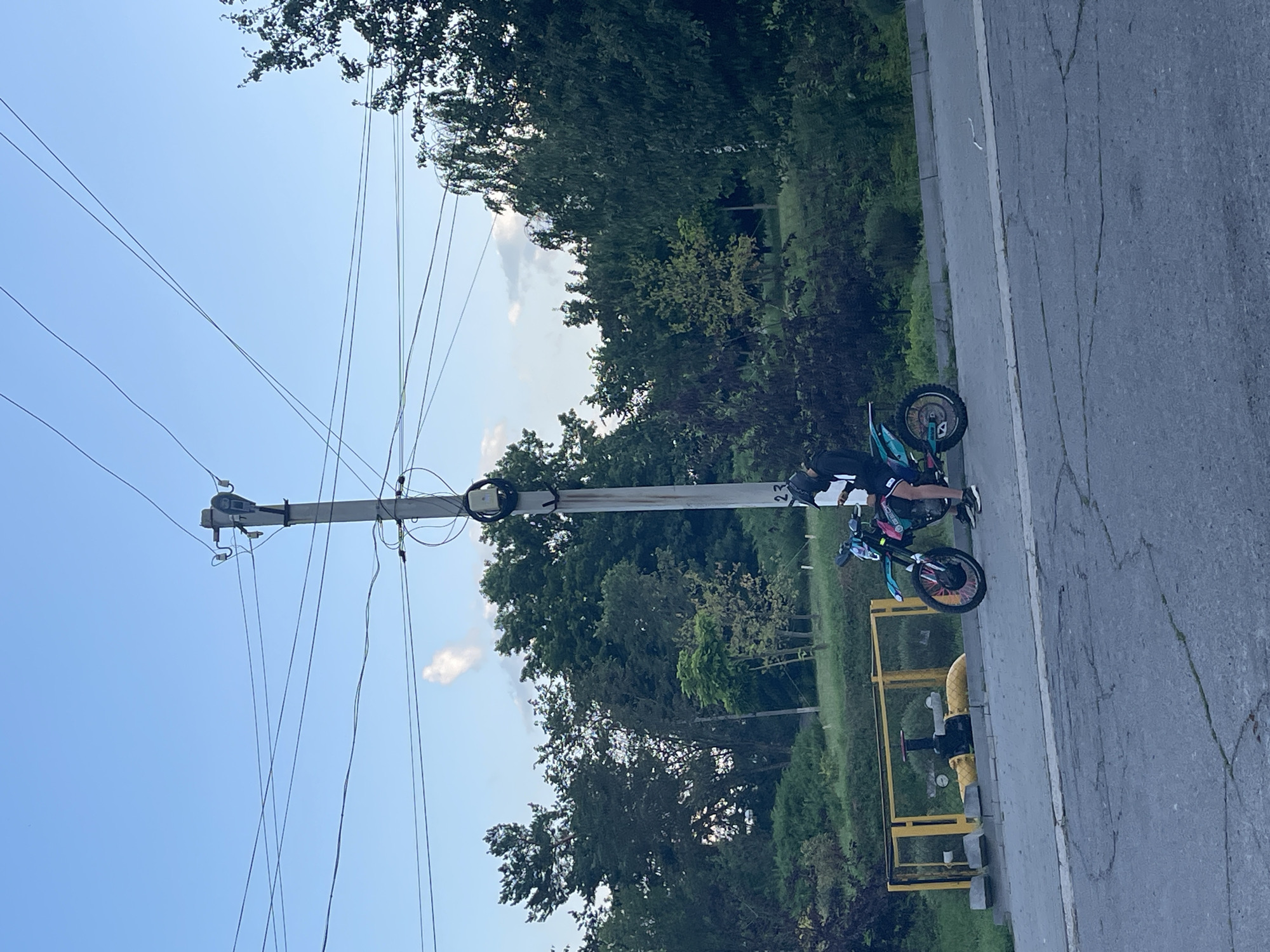 The image shows a person on a dirt bike parked on the side of a paved road. The person is wearing black shorts, a black and white patterned shirt, and black and white shoes. The dirt bike is teal and black with colorful accents on the wheels. The bike is leaning on its kickstand.
Behind the road, there is a grassy area with trees and bushes. A tall utility pole with several wires runs diagonally across the frame, stretching into the sky. The sky is clear and blue, with a few white clouds visible. A yellow gas pipe with a valve is located near the side of the road, behind the dirt bike.
The image appears to be taken outdoors during the daytime, likely in the late afternoon or early evening given the soft lighting. The setting suggests a rural or suburban area. There is no visible text in the image. The overall mood is relaxed and casual, with the subject appearing to take a break. The presence of the utility pole and the gas pipe suggests an area with infrastructure, possibly on the outskirts of a town.