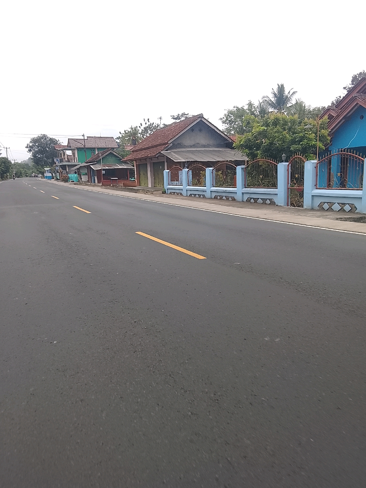 A paved road in Bangunsari, Indonesia, with a yellow line marking the lane, runs through a residential area. On the right side of the road, a row of houses with various roof colors and styles are visible, separated by a pale blue fence with ornate metalwork. Some houses have a vibrant blue facade, while others feature brown or gray roofs and walls. Green trees and vegetation are interspersed among the houses. On the left side of the road, more houses and some utility poles are present. The sky is overcast and bright. A motorcycle is seen in the distance on the left side of the road.