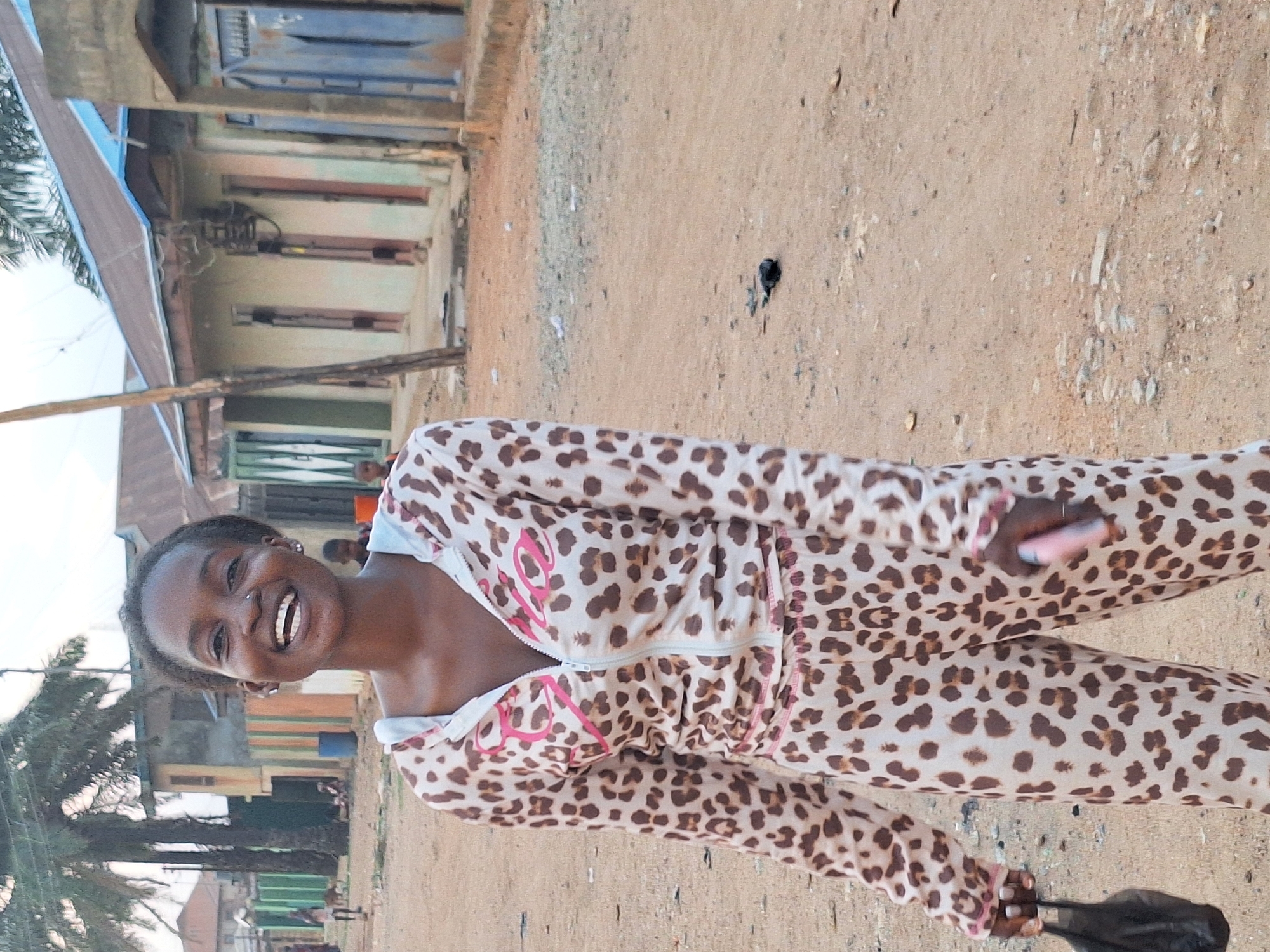 A young woman stands and smiles at the viewer, wearing a beige and brown leopard-print tracksuit with pink detailing. She holds a dark object in her left hand. The location appears to be Sabon Tasha, Nigeria, judging by the architecture and surroundings. The background includes buildings with a mix of concrete and corrugated metal roofing, along with a dirt ground and palm trees.