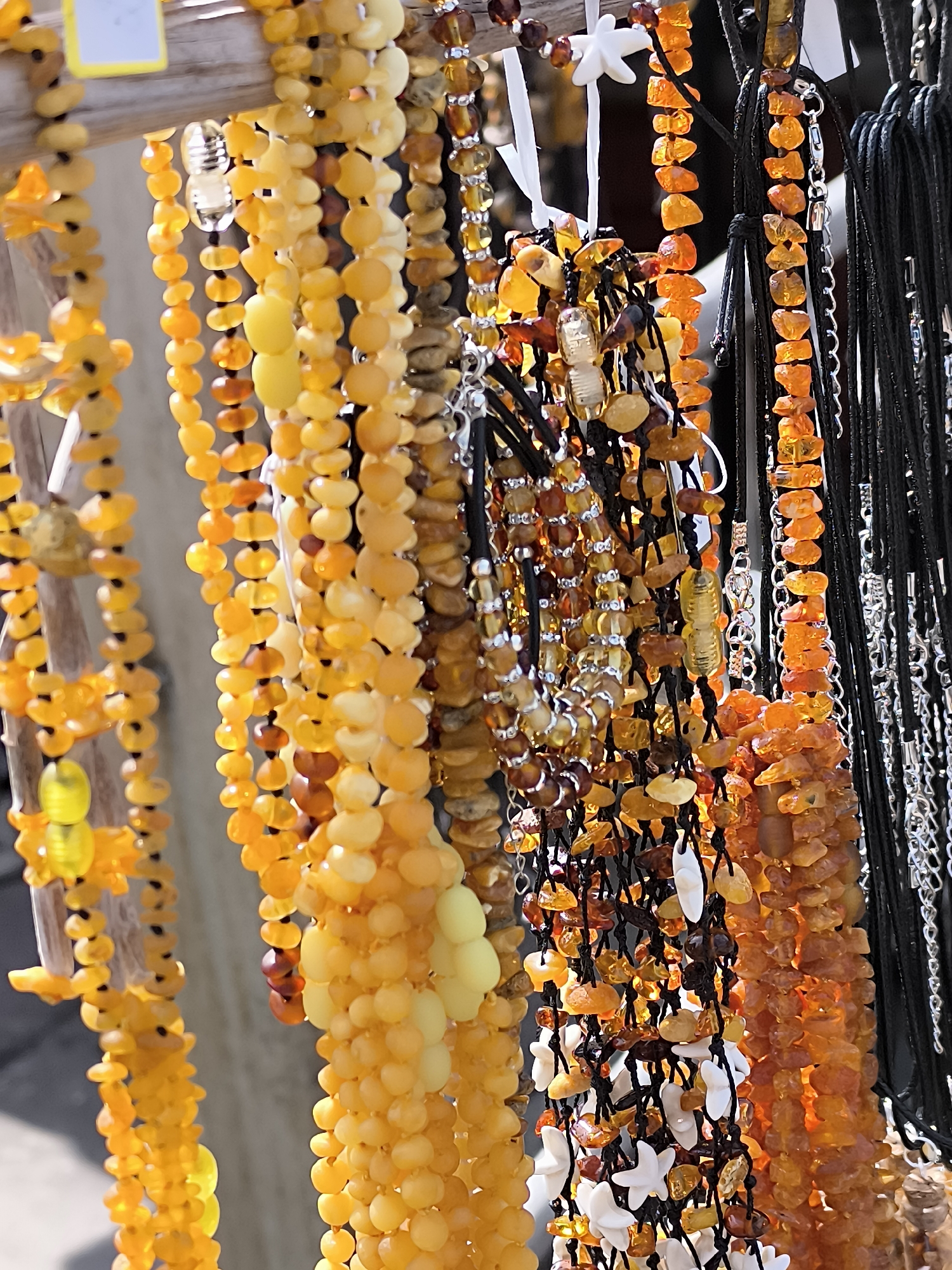 A close-up, vertical, eye-level shot captures a collection of amber necklaces displayed outdoors in Neringa, Lithuania. The necklaces, made from polished and raw amber beads in various shades of yellow, orange, and brown, hang from a wooden structure. Some necklaces are delicate strings of small, uniform beads, while others feature larger, irregularly shaped stones. Several necklaces are strung on black cords, some interspersed with small silver or white beads. One necklace on the far left is made of larger, translucent yellow beads. The background is out of focus, suggesting an outdoor market or street stall setting. The lighting indicates it is daytime, with sunlight illuminating the amber, making its colors vibrant. No people are visible in the frame.