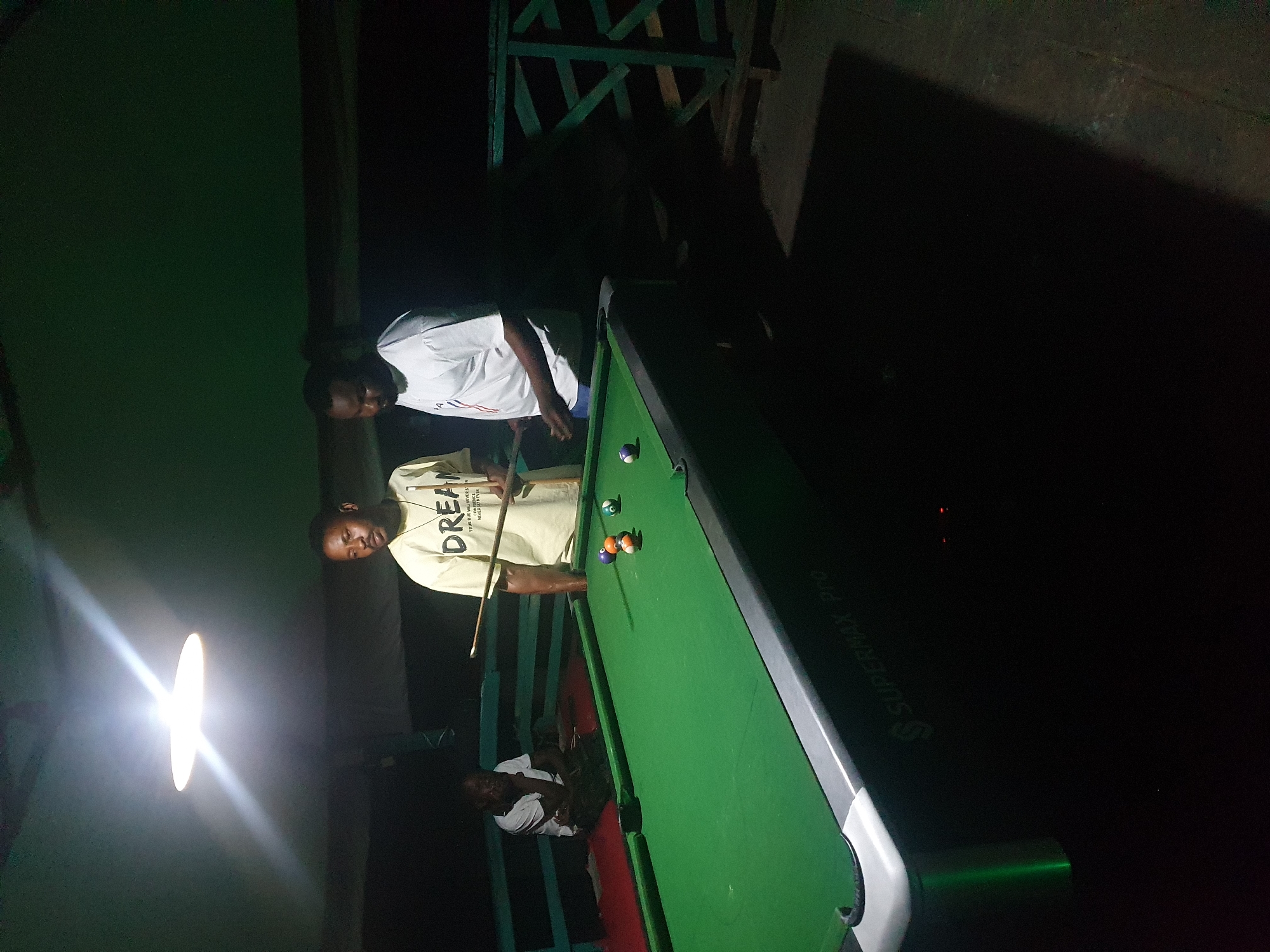 An outdoor, dimly lit billiards scene in Lokoja, Nigeria shows three individuals near a green pool table. Two men stand on the right side of the table. One, wearing a yellow t-shirt with "DREAM" printed on it, holds a pool cue, appearing to be lining up a shot. The second man, to his right, wears a white t-shirt and observes the game. A third individual sits on a bench to the left of the table, partially obscured by shadow. Several pool balls are scattered on the table, with one blue and white ball visible in the foreground left pocket. The table itself is a "SUPERMAX PRO" model. The environment suggests a casual setting, possibly a backyard or an outdoor recreational area, with a wooden railing visible in the background. The lighting originates from a single overhead lamp, casting long shadows.
