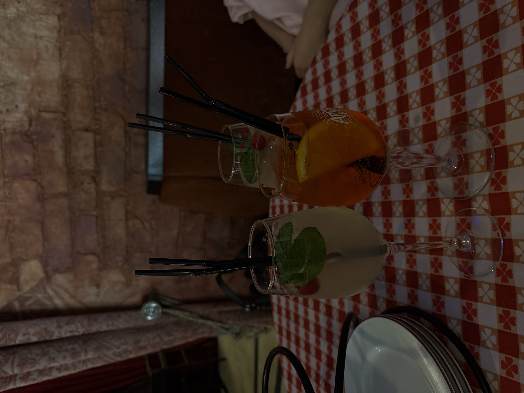 A close-up captures two drinks on a table with a red and white checkered tablecloth. The drink on the left is light-colored with mint leaves and a red fruit, likely a cocktail. The drink on the right is orange with a lemon slice and also includes mint leaves, possibly an Aperol Spritz. Both drinks have black straws. Two empty wine glasses are behind the drinks. A stack of white plates with red trim sits at the bottom of the frame. A blurred brick wall is visible on the left side of the image. The top of a person is seen in the upper center of the frame, wearing a light pink shirt. It's difficult to ascertain if the location is in Saint Petersburg, Russia without further information or context.