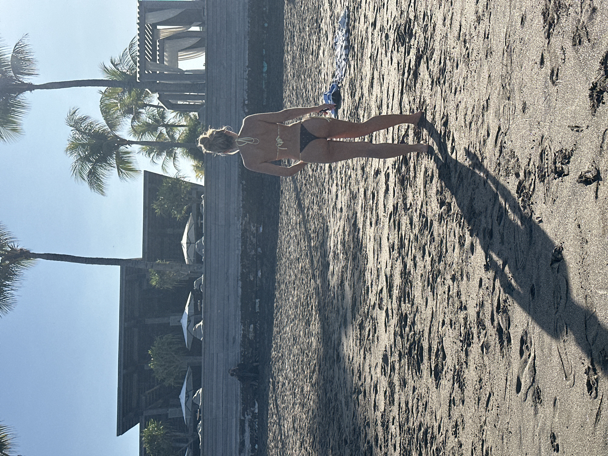 A woman stands on a sandy beach in front of a resort in North Kuta, Indonesia. She is wearing a black bikini and has her hair pulled up in a bun. The beach is covered in sand and there are footprints visible. In the background, there are palm trees, a building with a modern design, and some outdoor seating areas with umbrellas. The sky is clear and blue, and the sun is shining. The woman's shadow is cast on the sand in front of her. There is a towel and a pair of flip-flops on the sand to her right.