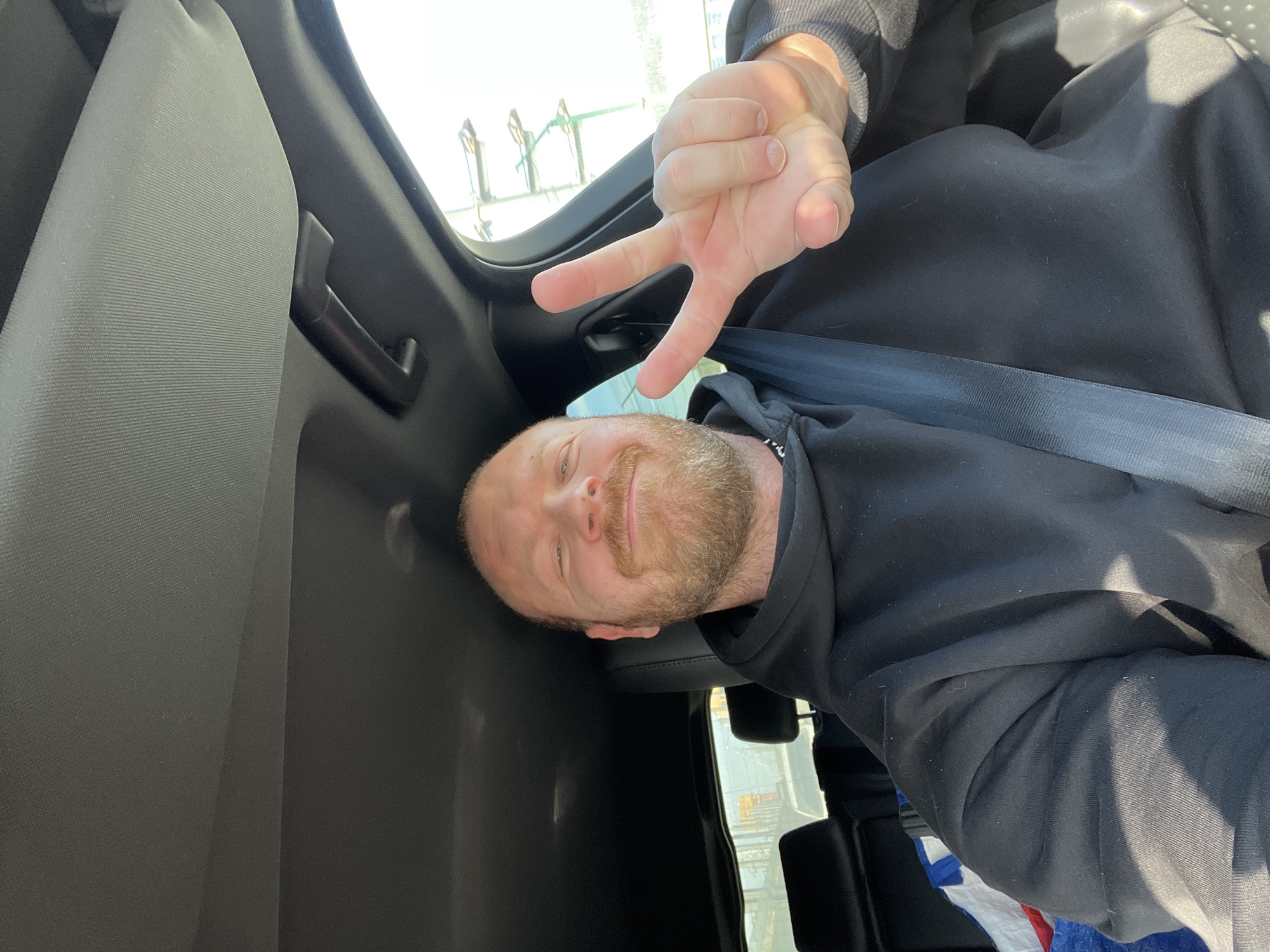 A male individual is seated in the driver's seat of a vehicle during daytime. He has a reddish-brown beard and short or bald hair, a dark stud piercing in his left nostril, and is smiling directly at the camera. He wears a black hooded sweatshirt and is secured by a black seatbelt across his chest and lap. His left hand is raised, forming a "peace" sign with the index and middle fingers extended. The vehicle's interior features a dark headliner, a light grey sun visor folded up on the passenger side, and a dark grab handle. Dark-colored rear seats and headrests are visible, with a multi-colored object, possibly a flag or blanket displaying red, white, and blue patterns, partially visible on the rear seat behind the driver. Through the vehicle's windows, a bright outdoor environment is visible, featuring a large, multi-tiered green or metallic structural element to the right of the vehicle. The precise location within the city of Chaikovsky, Russia, cannot be determined from the visual information provided.
