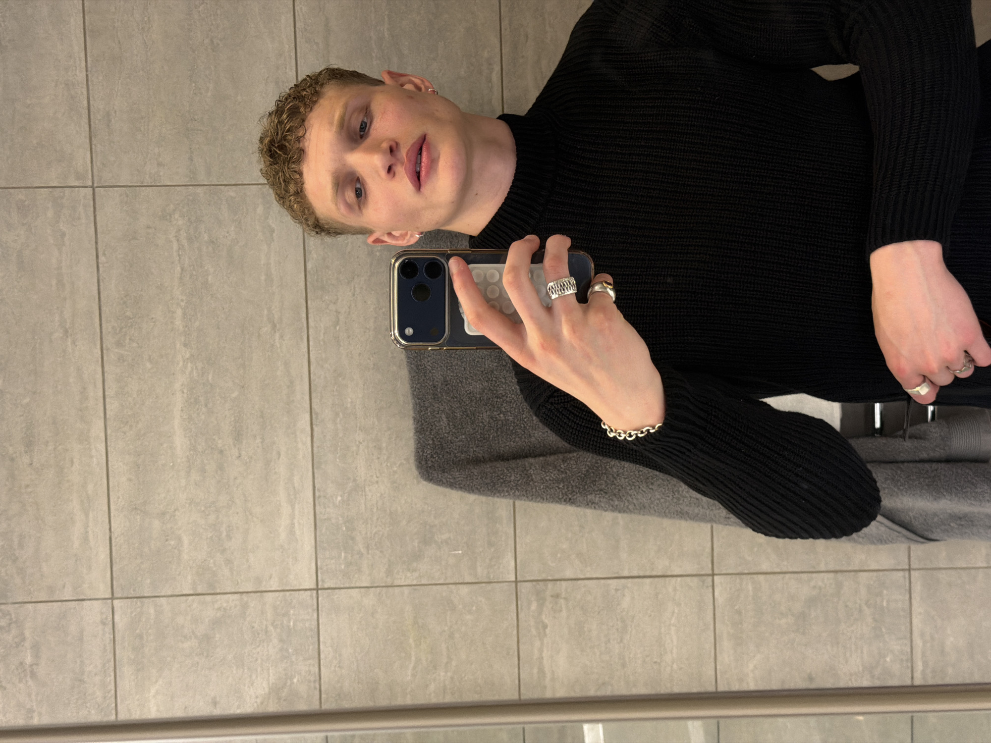 A young man with short, curly blonde hair is taking a selfie. He is wearing a black sweater and several rings, as well as a silver bracelet. He is holding a dark-colored smartphone with multiple camera lenses. The background consists of gray, square tiles, suggesting a bathroom or similar space. He is situated in a seemingly ordinary location. There is no clear indication that the picture was taken in London, UK.