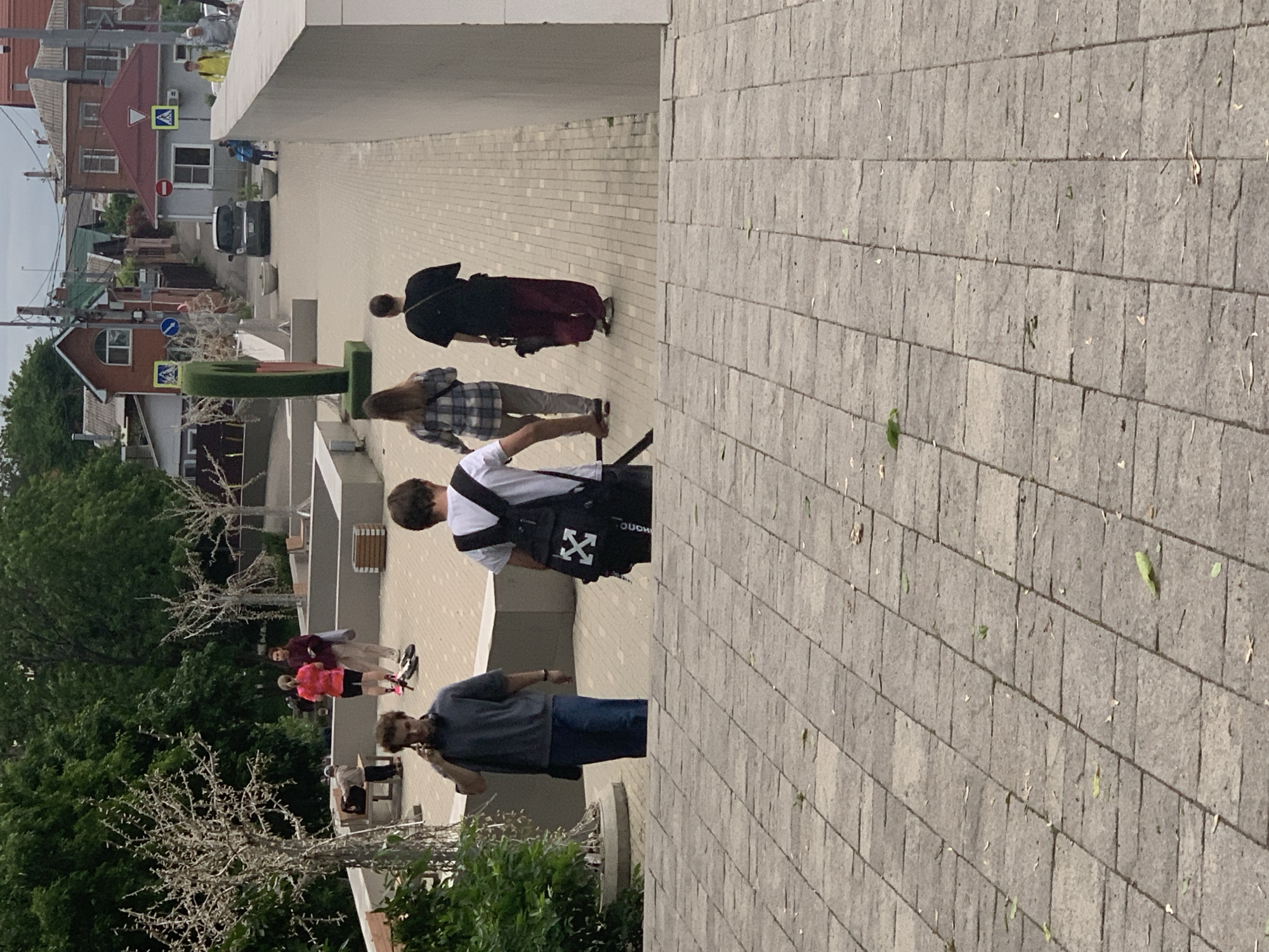 The image shows a street scene in Unknown, Unknown. Several people are walking on a paved path. A person in a grey shirt and jeans is holding a phone to their ear. Another person wears a white shirt and a black bag with a white "X" logo. Further down the path, there are two more people walking. Houses and trees are visible in the background.