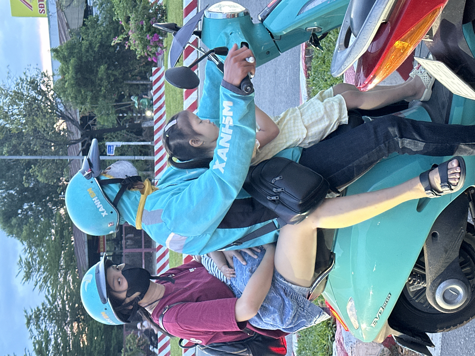 An adult wearing a blue helmet and a blue jacket is driving a teal scooter. They are wearing a dark-colored face mask and a yellow hood. A child with pigtails is sitting in front of the adult, wearing a light yellow checkered dress. Another adult, wearing a maroon t-shirt, black face mask, and blue jean shorts, is sitting behind the driver, holding a baby. The baby is wearing a striped onesie. The scooter has a black bag hanging from the handlebars and another black bag on the driver's left side. The scooter has a license plate. The background shows trees, a building with a sign, and a street with red and white striped barriers. The scene appears to be on a street in Đà Nẵng, Vietnam, given the style of the scooter, the clothing, and the presence of Vietnamese text on the driver's jacket, which appears to say "XANH".