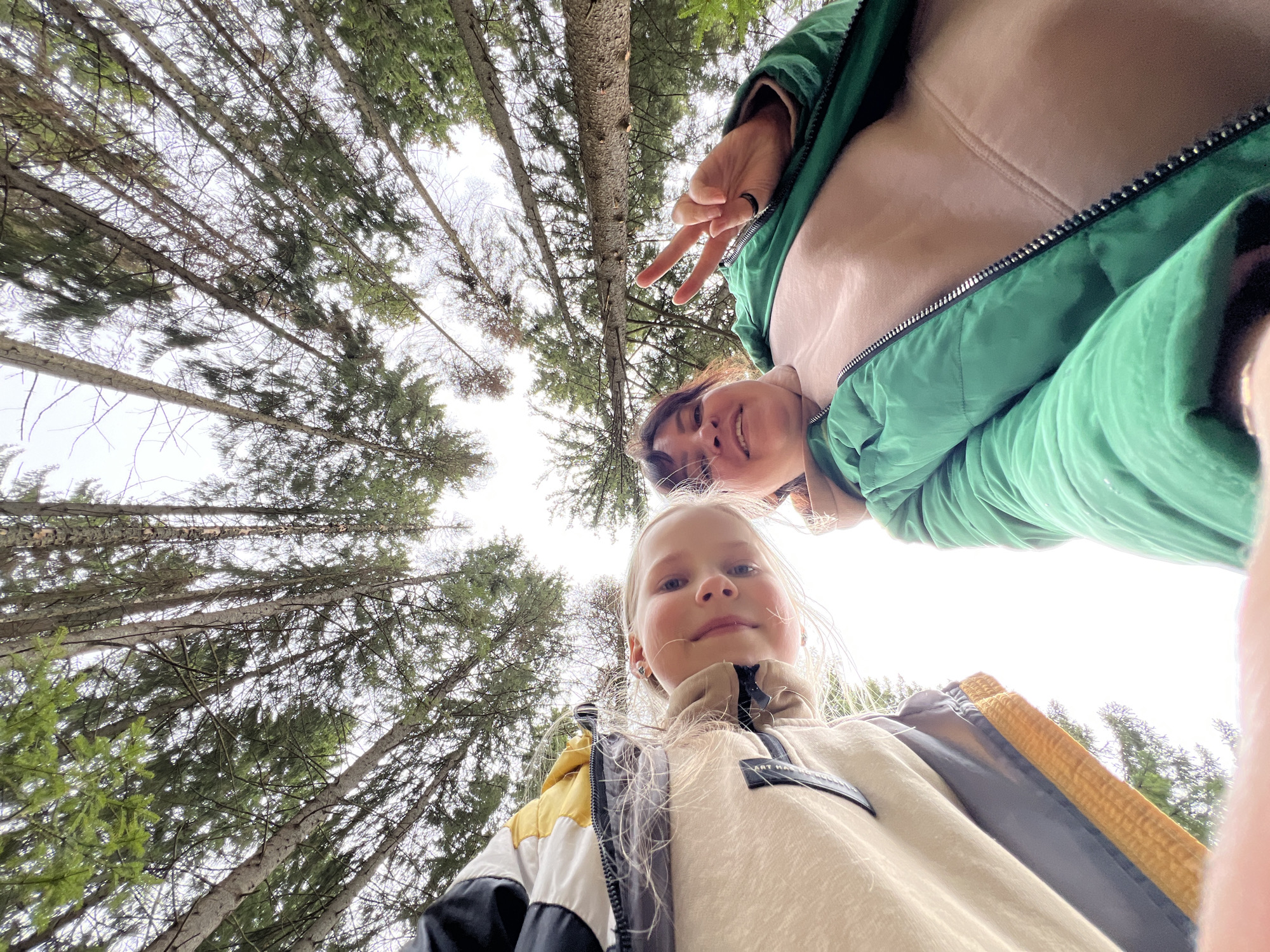 Two individuals, a girl and a woman, are positioned at the bottom of the frame, likely in Protasy, Russia, based on location. The vantage point is from a low angle, looking up. The girl is blonde. The woman has short, dark hair. The woman is giving a peace sign. Overhead, tall trees create a canopy, obscuring the sky.