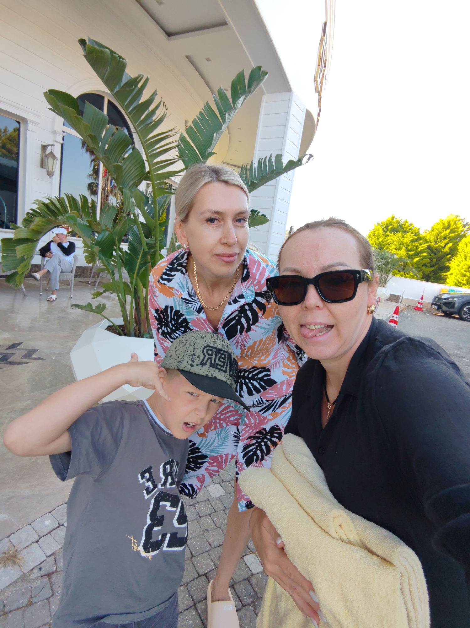 Three individuals, a woman, a younger boy, and another woman, are standing outdoors on a paved area with large paving stones in Alanya, Turkey. The boy is wearing a grey t-shirt with a number on it and a baseball cap. He is making a playful gesture with his hand near his face. The woman behind him is wearing a patterned shirt and a gold necklace. To her right, the other woman wears sunglasses and a dark shirt. She is sticking her tongue out. A light-colored towel is in her hands. Behind them is a white building with tropical foliage in a white planter. A man is sitting on a chair in the background. In the further background, trees, traffic cones, and a car are visible.
