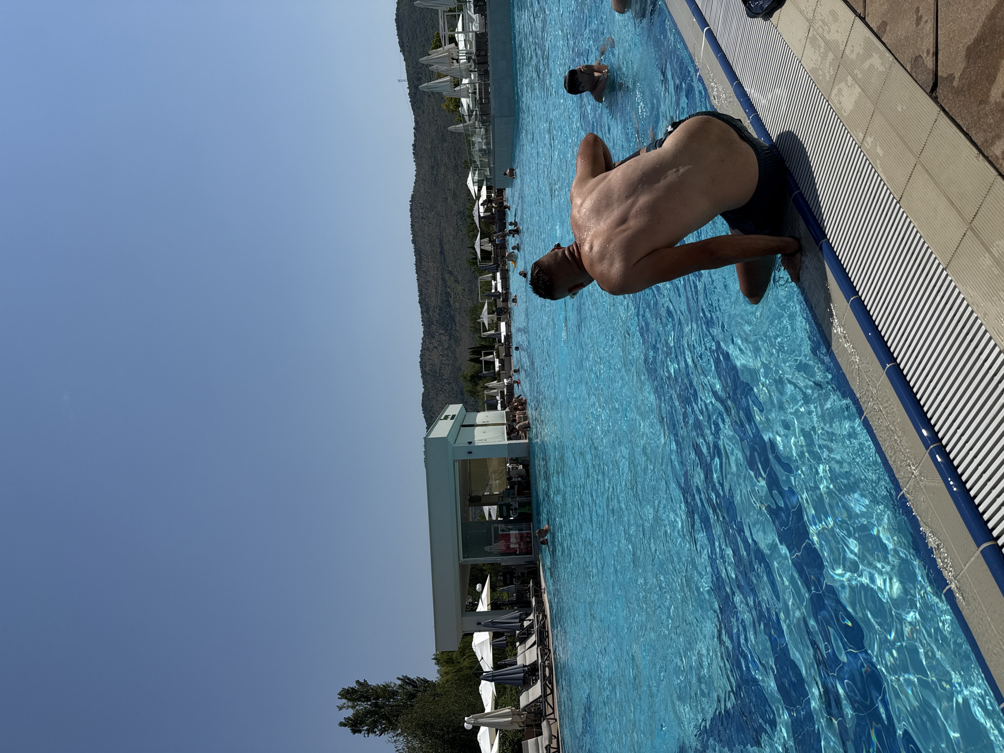 In an outdoor swimming pool on a clear day, a man in dark swim trunks sits on the edge of the pool, his back to the camera. His torso is wet and appears tanned. To his right and further in the pool, a young boy, also in swim trunks, splashes water with his hands near his face. In the background, several other people are visible in the water. Beyond the pool, there are lounge chairs with umbrellas, a modern white building with a bar area and patrons, and a lush, tree-covered hillside under a clear blue sky. The pool water is a vibrant turquoise, with reflections of sunlight creating patterns on the surface. The edge of the pool is lined with striped gray and blue tiles.