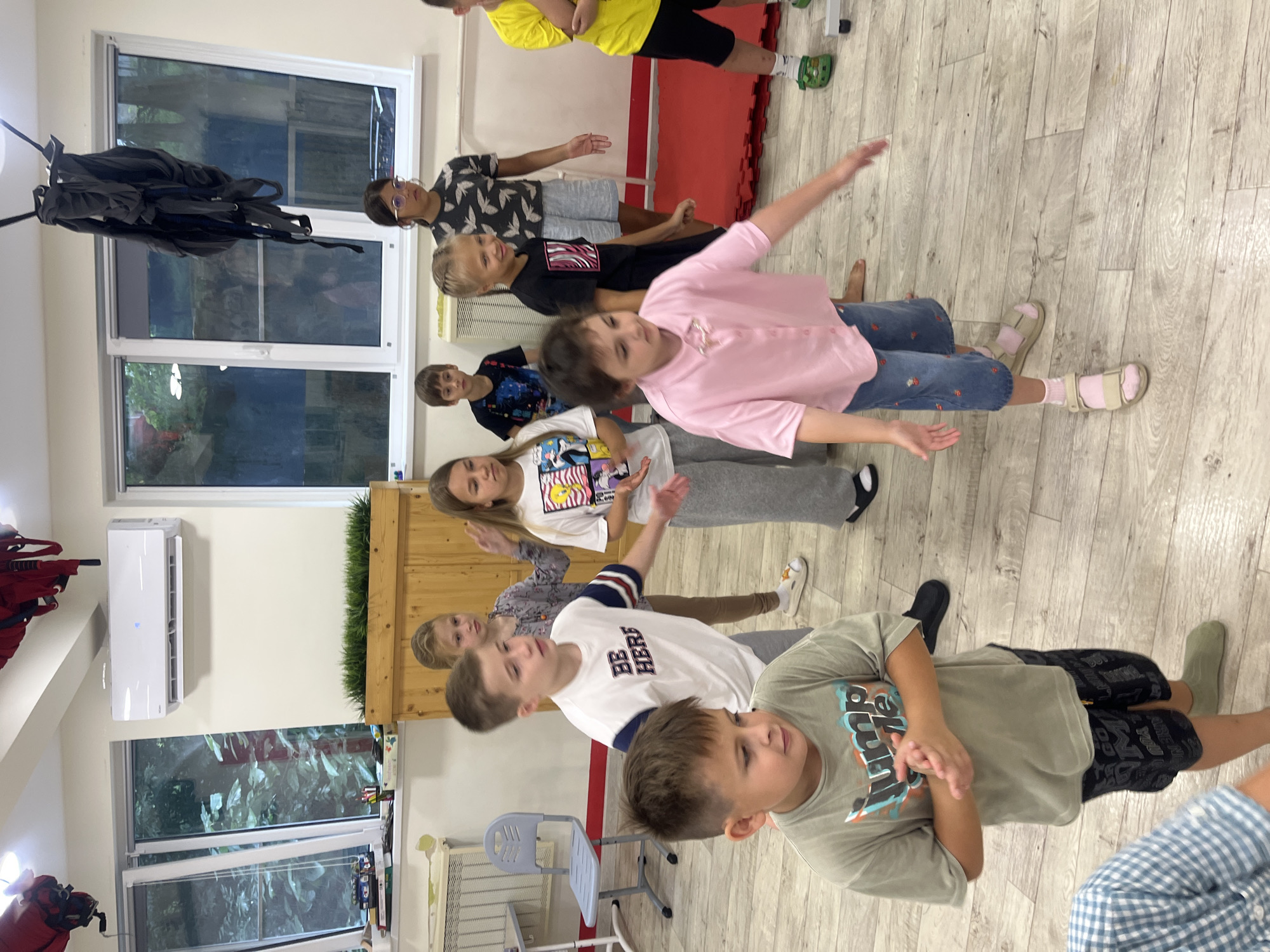 The image depicts a group of children in a room, possibly a studio or gymnasium, with wooden floors and light-colored walls. An air conditioning unit is mounted high on one wall. Several windows offer a view of greenery, suggesting an outdoor environment. The children are dressed in casual clothing, and some appear to be engaged in an activity or instruction.
In the foreground, a young boy in a green t-shirt and patterned shorts is looking down. To his right, a girl in a pink shirt and denim shorts is actively engaged, with her arms outstretched. Behind them, other children are visible in various poses, some looking towards the front, others interacting amongst themselves. A chair is positioned to the left of the frame, and red mats are visible on the floor in the background. The overall atmosphere suggests a lively and active setting for the children.
The image is from Sochi, Russia, specifically from the "Sochi Sports Club" (Сочинский спортивный клуб), located at Novorossiyskaya Street, 18.