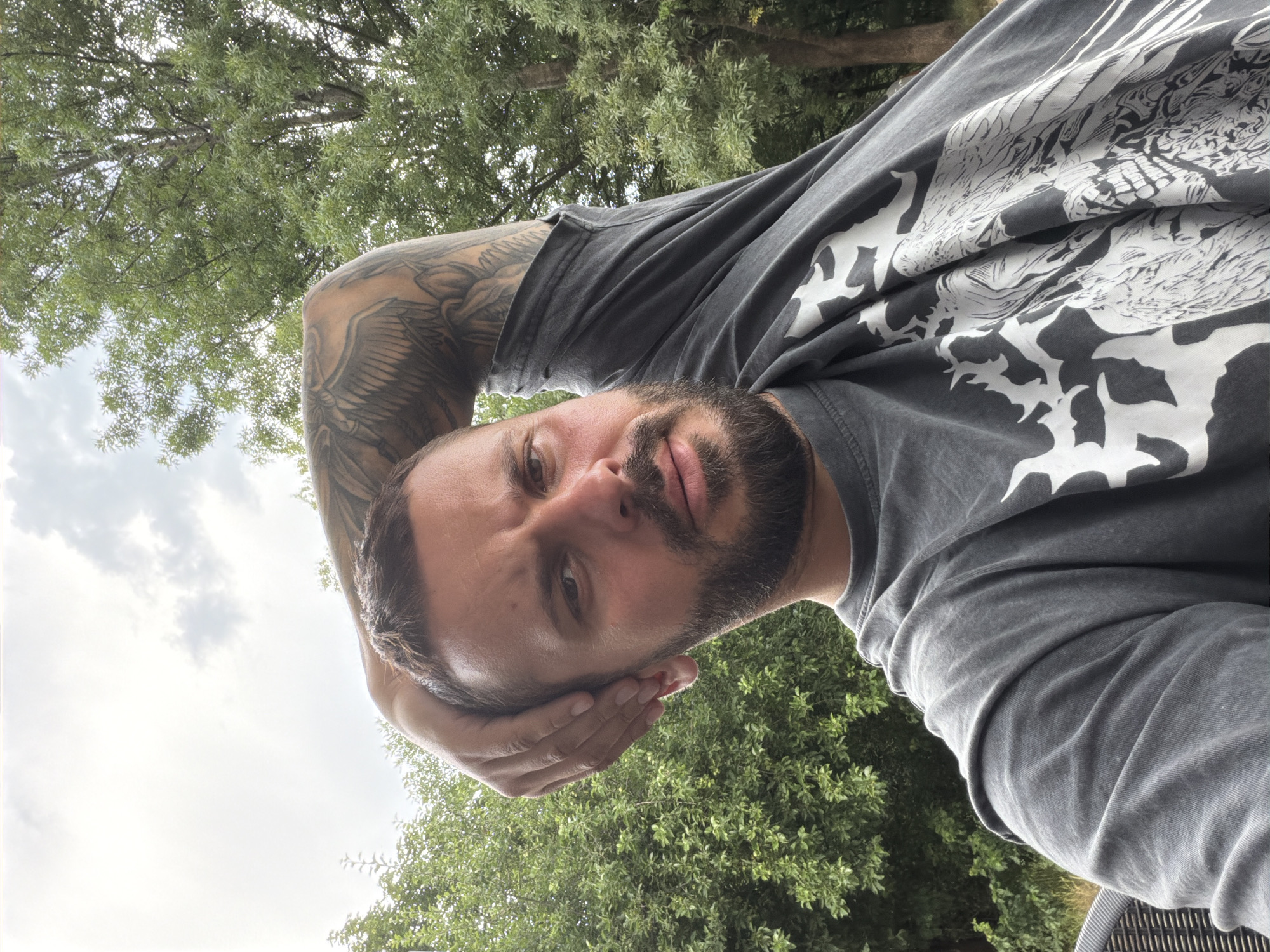 A man with a beard and tattoos on his arm is seen in a selfie. He is wearing a grey t-shirt with a white graphic design. His left arm is raised and resting on the back of his head. He is outdoors, with trees and a cloudy sky in the background. The scene appears to be a park or garden area, possibly in Sofia, Bulgaria, given the context.