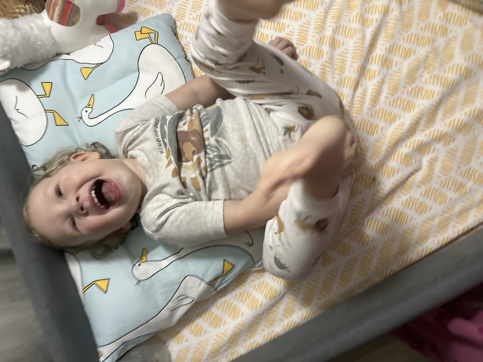 A toddler in a patterned pajama set is lying on their back on a bed, with their head on a blue pillow with white swan designs. The toddler is smiling widely with their mouth open. A stuffed toy is partially visible in the upper left corner of the image. The bedsheets have a yellow and white geometric pattern. The image is taken from a high-angle perspective. The location cannot be identified as Votkinsk, Russia from the provided image.