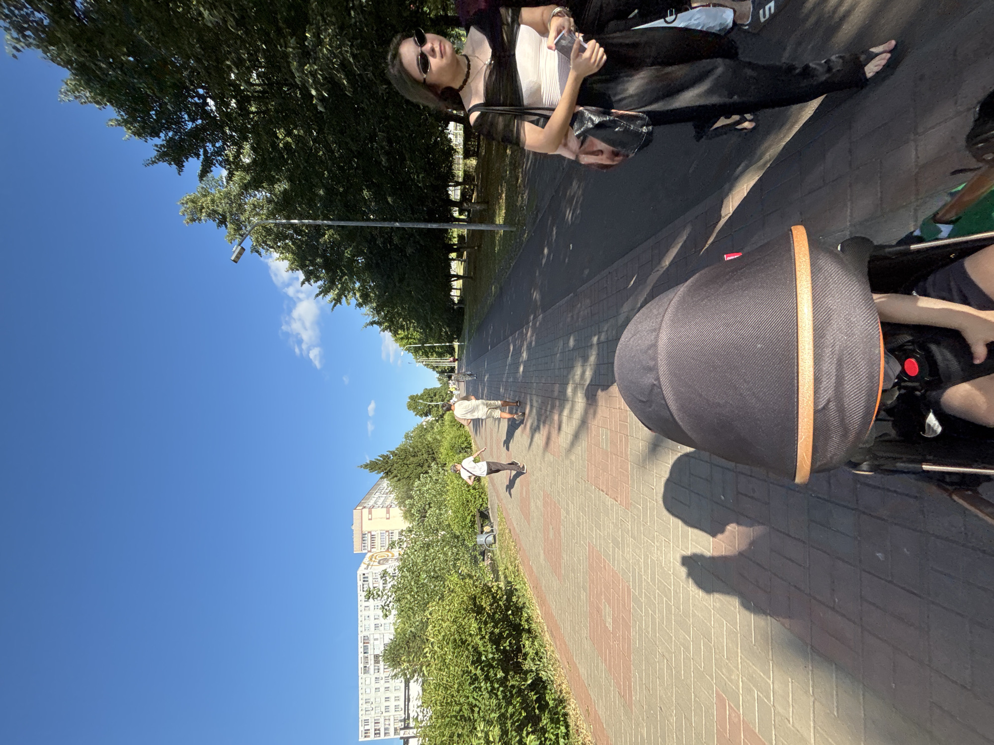 The image captures a sunny day on a paved walkway in Naberezhnye Chelny, Russia. The walkway is constructed of red and grey bricks, bordered by lush green bushes on the left and a line of trees and a paved road on the right. In the background, multi-story buildings are visible, typical of urban residential areas.
Several individuals are present. In the foreground, a baby stroller is positioned, suggesting a person pushing it is taking the photo from a first-person perspective. Further down the walkway, two men are walking away from the camera, one in a white t-shirt and dark pants, the other in a light t-shirt and shorts. On the right side of the image, a young woman with dark hair, wearing sunglasses, a black and white striped top, and dark pants, is walking towards the camera. She is carrying a bag over her shoulder and holding a phone. Another person is partially visible behind her. The sky is a clear, bright blue with a few wispy clouds. Lampposts are spaced along the right side of the walkway. The overall impression is a casual stroll in a city park or promenade.