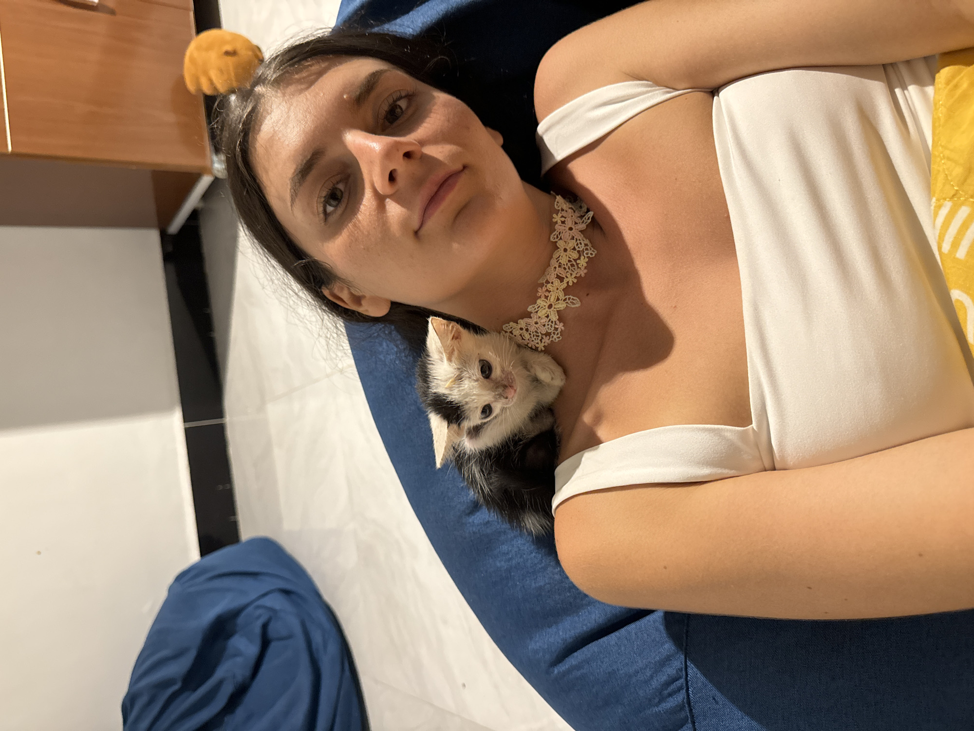A woman is lying down on a blue beanbag chair. A black and white kitten is perched on her left shoulder. The woman is wearing a white tank top and a delicate floral choker necklace. Behind her, a wooden cabinet and a white wall are visible. The flooring appears to be tiled. The scene is set indoors, likely in a residential area or hotel room in Mui Ne, Vietnam.