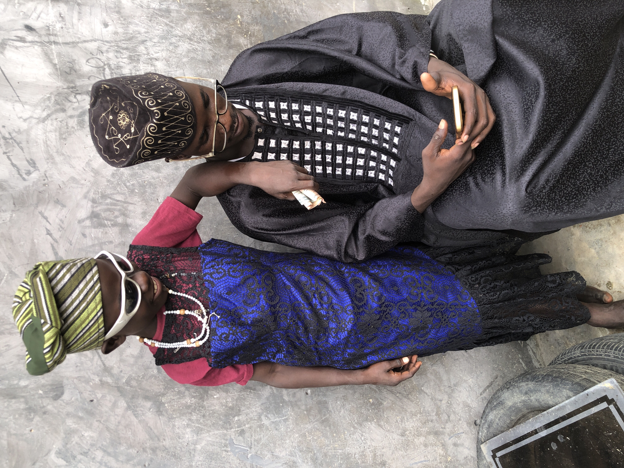 In the city of Ado, Nigeria, a child in a vibrant green and white striped cap, white sunglasses, and a pearl necklace stands to the left of a seated man. The child wears a maroon shirt under a blue lace dress with a black ruffled hem. The child's left arm rests on the seated man's shoulder, and they are holding what appears to be cash. The man is dressed in a black traditional garment with white embroidered squares on the chest and wears a black cap with gold embroidery and gold-rimmed glasses. He is looking down at his phone, which he holds in his hands. The background is a textured, weathered gray wall. To the left of the seated man, stacked tires are visible.