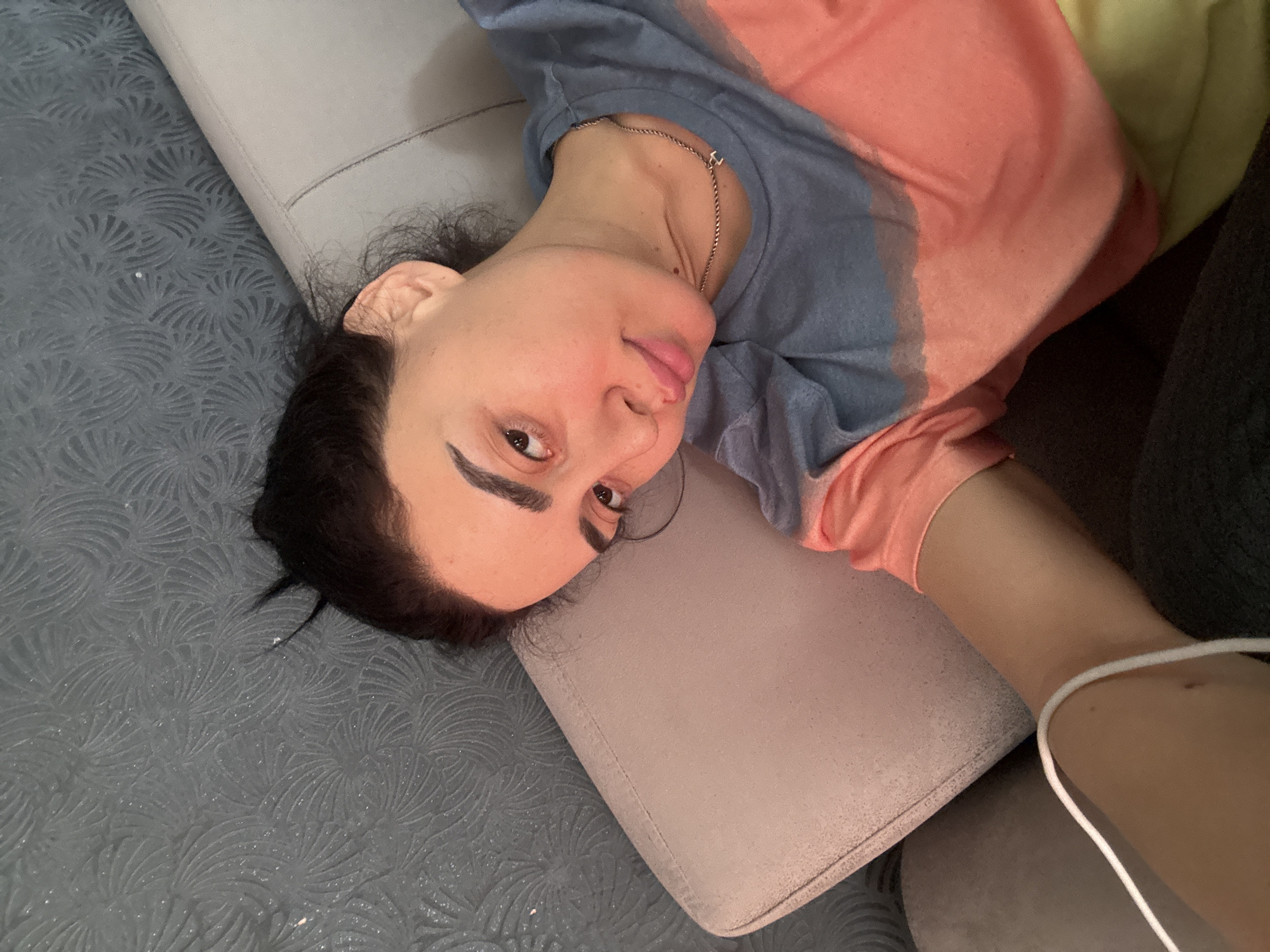 A woman lies on a gray sofa. She wears a blue and peach t-shirt. She is in Tbilisi, Georgia. The sofa has a patterned covering.