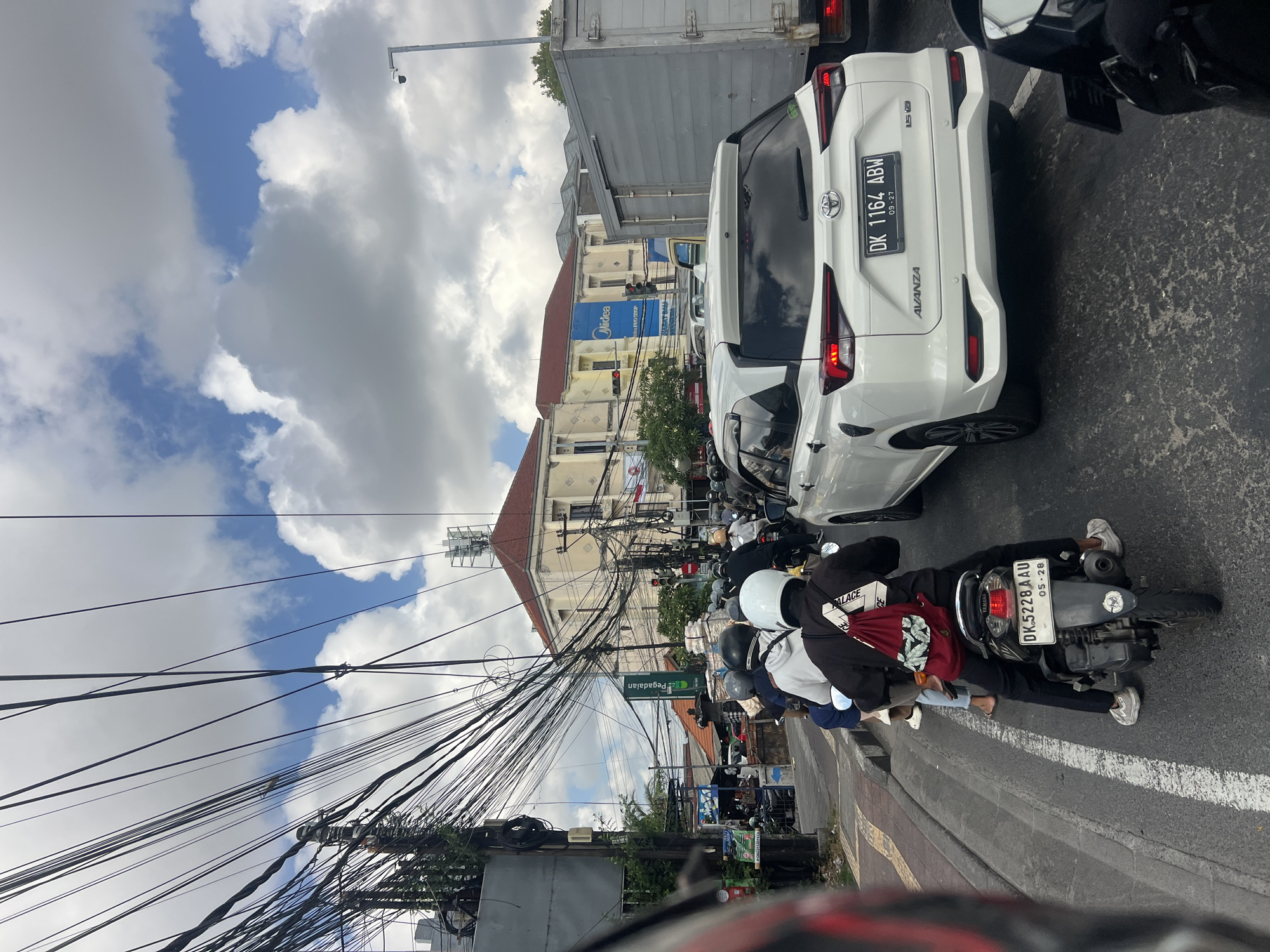 The image depicts a congested street scene in Denpasar, Indonesia, likely during daylight hours given the bright sky. A multitude of motorcycles and a white car are stopped in traffic, indicating a busy urban environment. The foreground is dominated by the back of a motorcycle with a rider wearing a dark jacket and a white helmet, carrying a red backpack. Another motorcycle and a white car are visible ahead. The background shows a densely packed cityscape with buildings, including one with a red-tiled roof and a blue sign that reads "Midea". Overhead, a chaotic network of electrical wires crisscrosses the sky, a common sight in many Indonesian cities. The sky is partly cloudy with patches of blue. The presence of traffic lights and multiple vehicles suggests a significant road. The license plates on the vehicles are visible, with one on the white car reading "DK 1164 ABW". The overall impression is one of urban hustle and bustle.