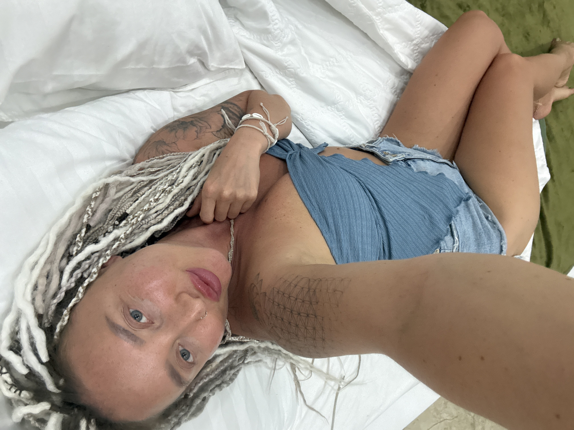 This media file shows a woman lying on a bed. She has long, light-colored dreadlocks and several tattoos on her arms. She is wearing a blue top and denim shorts. Her eyes are open and she is looking at the camera. The bed has white sheets and pillows, and a green blanket or rug is visible to the right. The scene appears to be indoors, possibly in a bedroom. There are no visible signs indicating the time of day or weather. The woman's expression is neutral. No text is visible in the media file. The location context is Bac Nha Trang Ward, Vietnam.