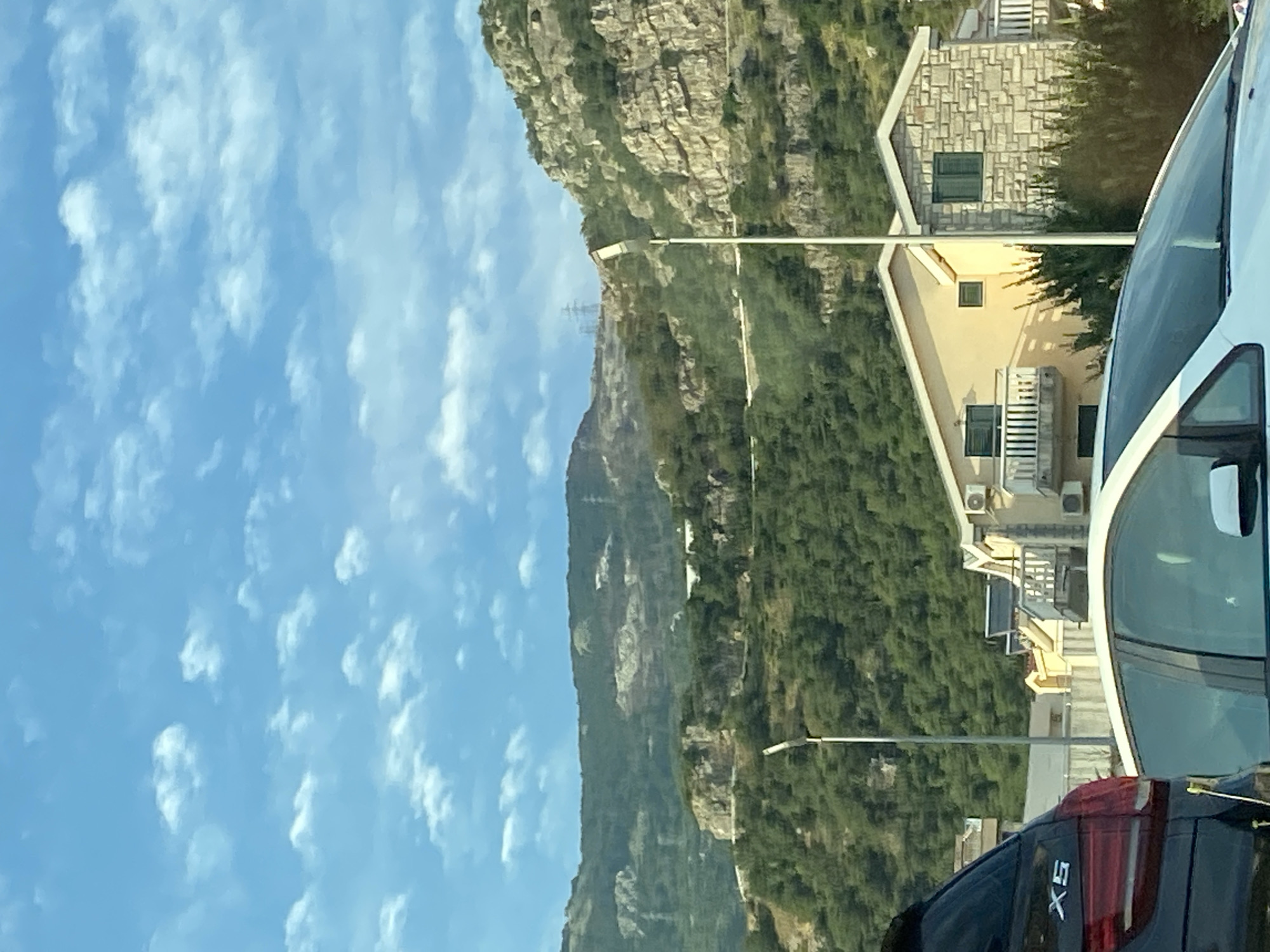 The image depicts a view of a mountainside with lush green vegetation and rocky outcrops. The sky is a clear blue with scattered white clouds. In the foreground, a portion of a white car is visible on the right, and the rear of a black car with the "X5" logo is on the bottom right. Further back, there are buildings with light-colored facades and tiled roofs, nestled into the hillside. One building has a facade covered in stone tiles. Balconies and air conditioning units are visible on the structures. A street light stands on the left side of the frame. The scene appears to be a residential area built on a steep slope, overlooking a landscape with natural and man-made elements. The lighting suggests it is daytime, likely sunny given the clear sky and the shadows cast by the buildings. The overall impression is of a scenic, somewhat rugged coastal environment. The location context is Budva, Montenegro.