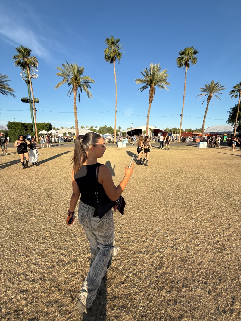 A woman walks through a dry grassy field, likely at the Coachella Valley Music and Arts Festival in Indio, California. She is wearing a black tank top, patterned pants, and a black shoulder bag. She is holding a phone in her right hand. Palm trees line the background of the image, and tents with people walking in them can be seen in the distance. The sky is blue with scattered clouds.