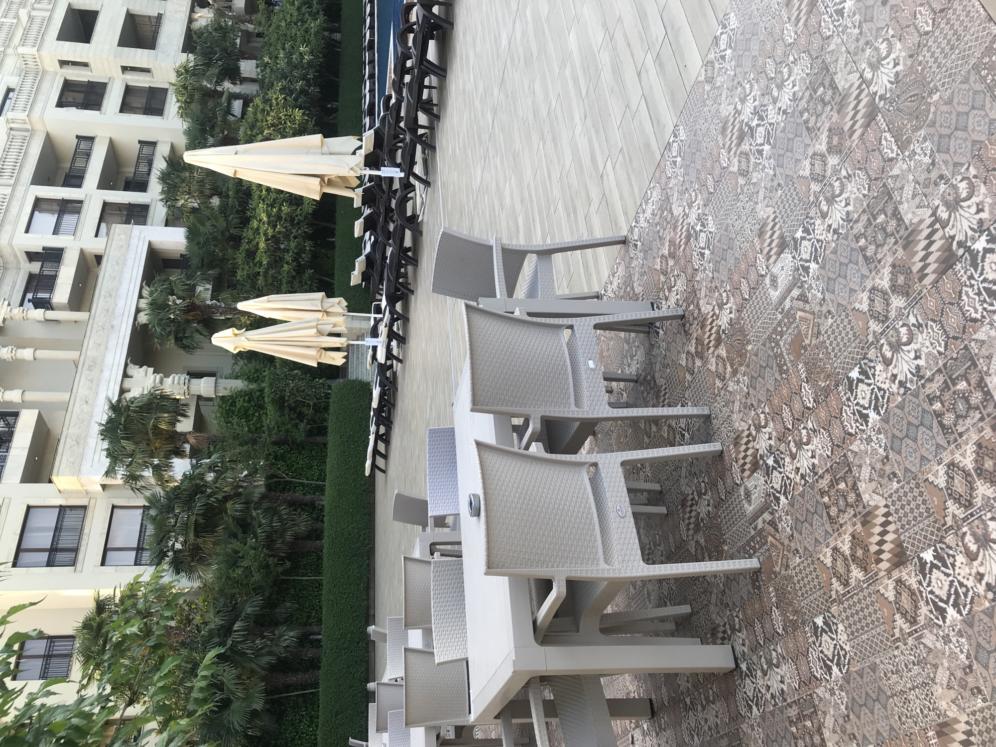 The image depicts an outdoor seating area adjacent to a swimming pool and a large hotel building in Varna, Bulgaria. The foreground features several white plastic tables and chairs, arranged on a patterned tiled floor. In the middle ground, there are rows of unoccupied lounge chairs facing a blue swimming pool. Further back, several closed, light-colored patio umbrellas are visible. The background is dominated by a multi-story hotel with beige walls and dark-trimmed windows and balconies, surrounded by lush green trees and manicured hedges. The overall scene suggests a resort or hotel setting, possibly during daylight hours as indicated by the lighting and the closed umbrellas. There are no people visible in the image.