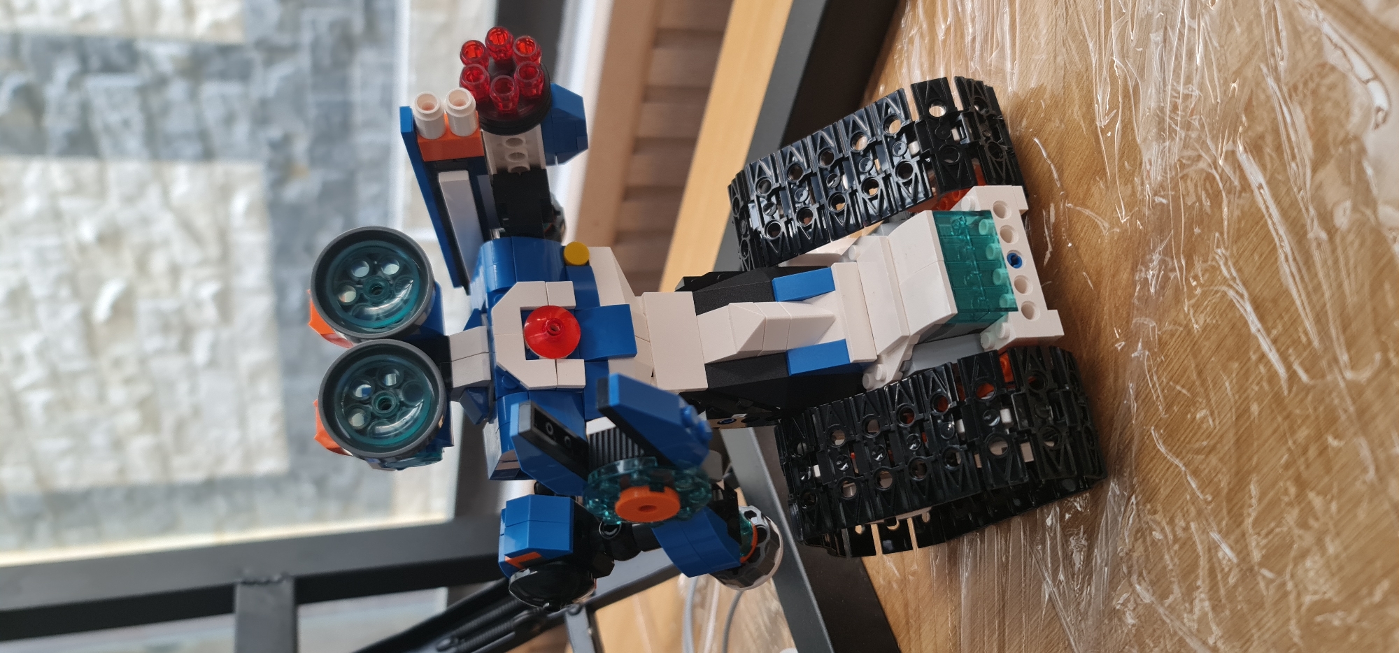 A detailed photograph shows a Lego construction toy, predominantly blue, white, and black, with orange and red accents. The toy is a vehicle-like structure with tracked wheels and what appears to be a missile launcher on top. The backdrop includes a window with a view of an indistinct urban area and a wooden surface covered with plastic wrap. The image is taken from a high angle, looking down on the toy. Đà Nẵng, Vietnam, is not identifiable within the photo.