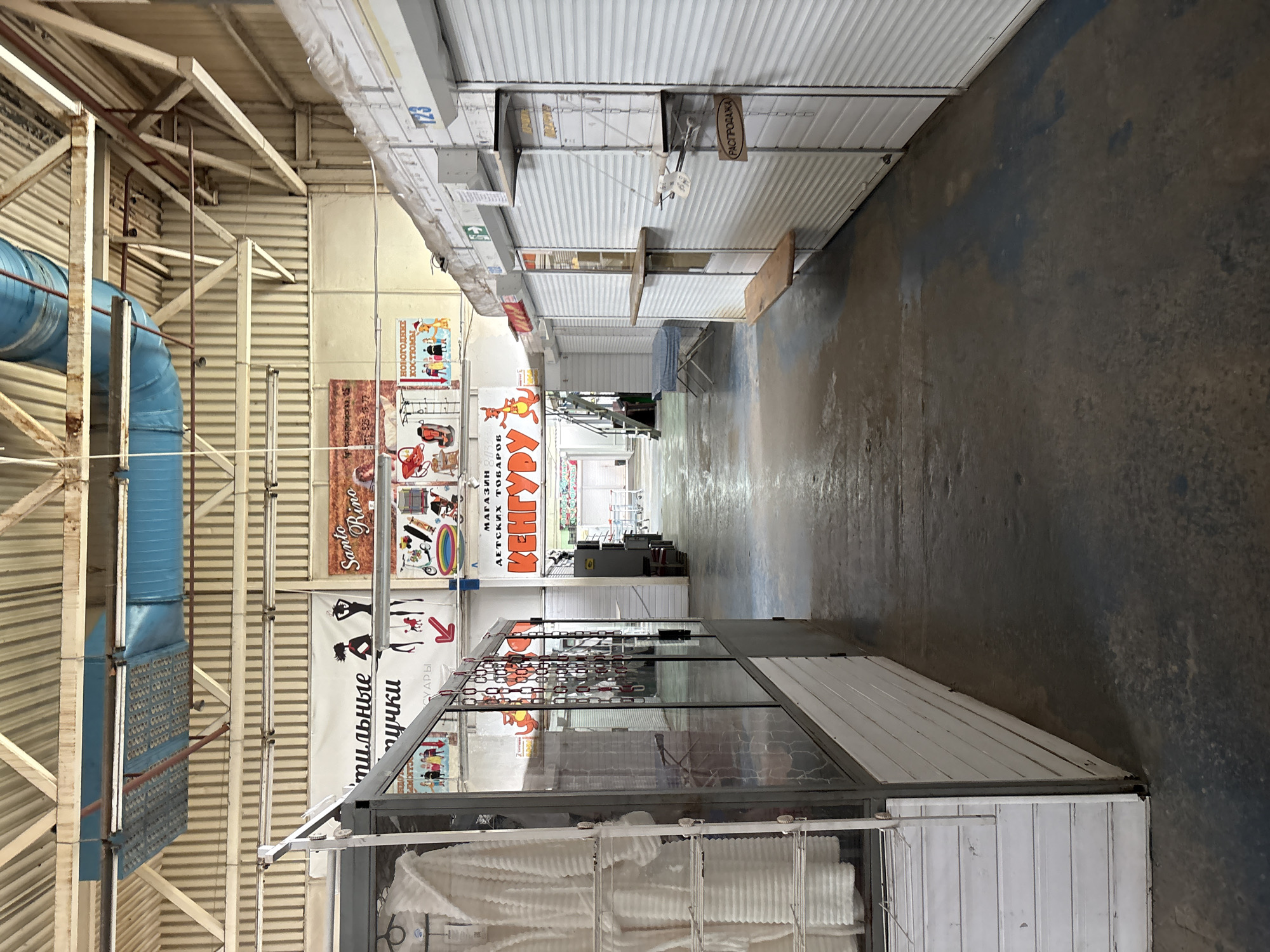 The image depicts the interior of a market or shopping center in Pastavy, Belarus. The scene is dominated by rows of closed stalls with white metal roll-up doors, suggesting a commercial environment that is either closed or in the process of closing for the day. In the foreground on the left, a stall displays white terrycloth robes on hangers behind a glass partition. Further down the aisle, the perspective leads to a more open area with visible shopping carts and a staircase. Overhead, industrial lighting fixtures and ventilation ducts are visible against the ribbed metal ceiling and supporting beams. Several signs with Cyrillic text are present, including one that reads "Santo Rino" and another with a depiction of a kangaroo and the text "ДЕТСКИХ ТОВАРОВ" (children's goods). Another sign, partially obscured, appears to indicate "РАСПРОДАЖА" (sale). The floor is a worn concrete surface, with some damp patches. The overall impression is of a functional, albeit somewhat aged, retail space.
